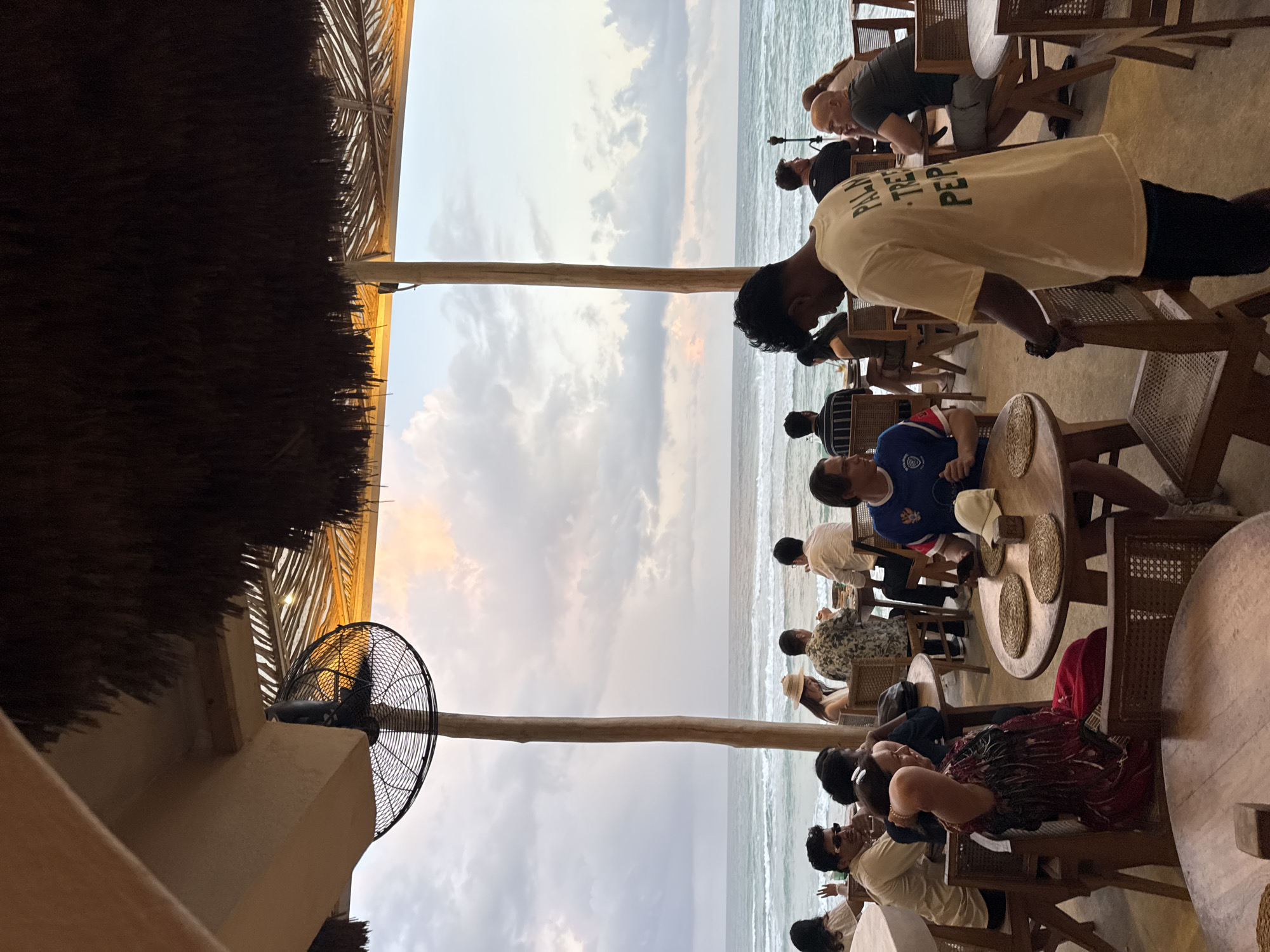 This image depicts an outdoor dining area with a view of the ocean. A thatched roof structure supported by wooden poles provides shade. In the foreground, patrons are seated at wooden tables, some eating and some conversing. A large industrial fan is mounted on a pole overhead. The ocean with gentle waves is visible in the background under a cloudy sky with hints of sunset colors. The scene appears to be at a restaurant or cafe located on the coast in Ahangama, Sri Lanka, given the architectural style and natural surroundings.