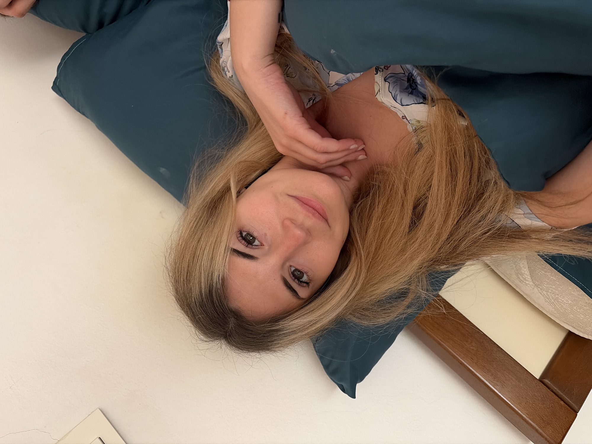 A woman with blonde hair and light skin lies on a dark blue pillow, partially covered by a matching blanket. She is wearing a floral-patterned top. Her right hand is near her neck. The background includes a white surface and a wooden frame. The location is unknown and cannot be determined from the visual information provided.