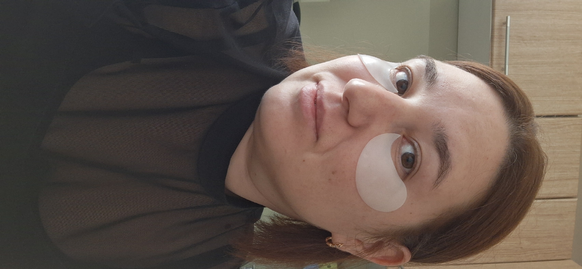 A woman with reddish-brown hair and fair skin is positioned facing the camera, angled slightly to the right. She has eye patches under both eyes. She is wearing a black, sheer top. Her left ear has a gold earring.
The background includes a beige wall, and a light brown wooden cabinet. The location of the photo is Chaikovsky, Russia.