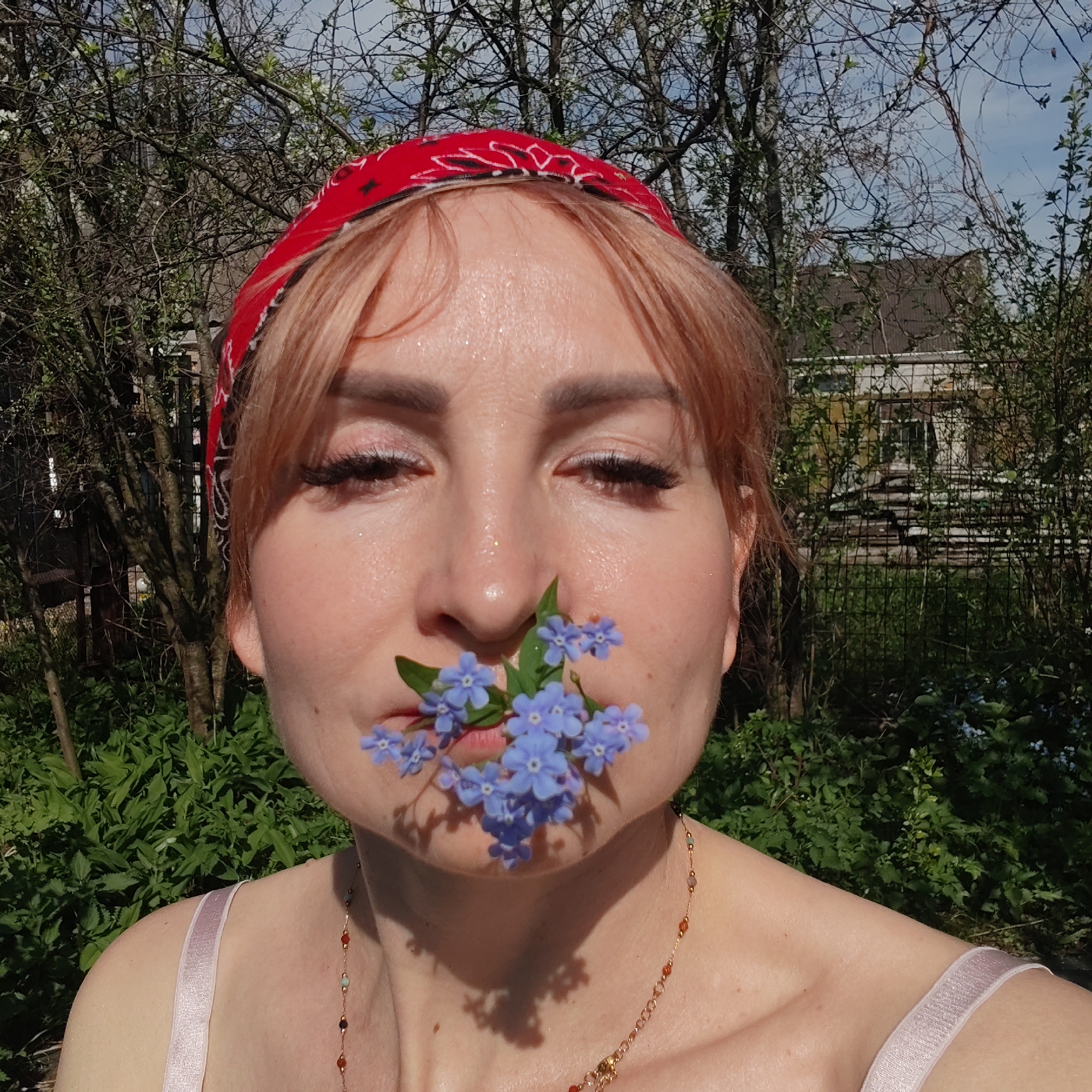 A woman is centered in the frame, holding a cluster of small, blue flowers between her nose and upper lip. She wears a red bandana with a black and white pattern on her head and a light-colored sleeveless top. A delicate necklace with multiple colored beads is visible around her neck.
Her skin appears slightly shiny, possibly due to sweat or moisturizer. Her eyes are partially closed, and she has eyelashes with makeup. Her hair is light red.
The background features greenery and trees, suggesting an outdoor setting. A structure resembling a building can be seen in the distance. It is unlikely that I can know where this image was taken without additional context.