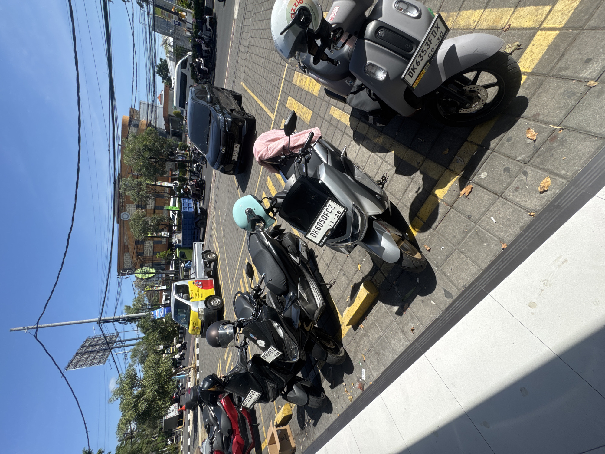 In Kuta, Indonesia, on a sunlit day with a clear blue sky, a row of parked motorcycles occupies a paved area marked with yellow parking lines. The foreground features a white tiled walkway casting a diagonal shadow. Among the motorcycles, a light blue helmet rests on one, and a pink fabric is draped over another. Several of the motorcycles display license plates, including one that reads "DK6050FCZ". In the background, a yellow and silver truck with "maxim" branding is parked, along with a black SUV and other vehicles on a road. Buildings, trees, and a tall utility pole with overhead wires are visible further in the distance.