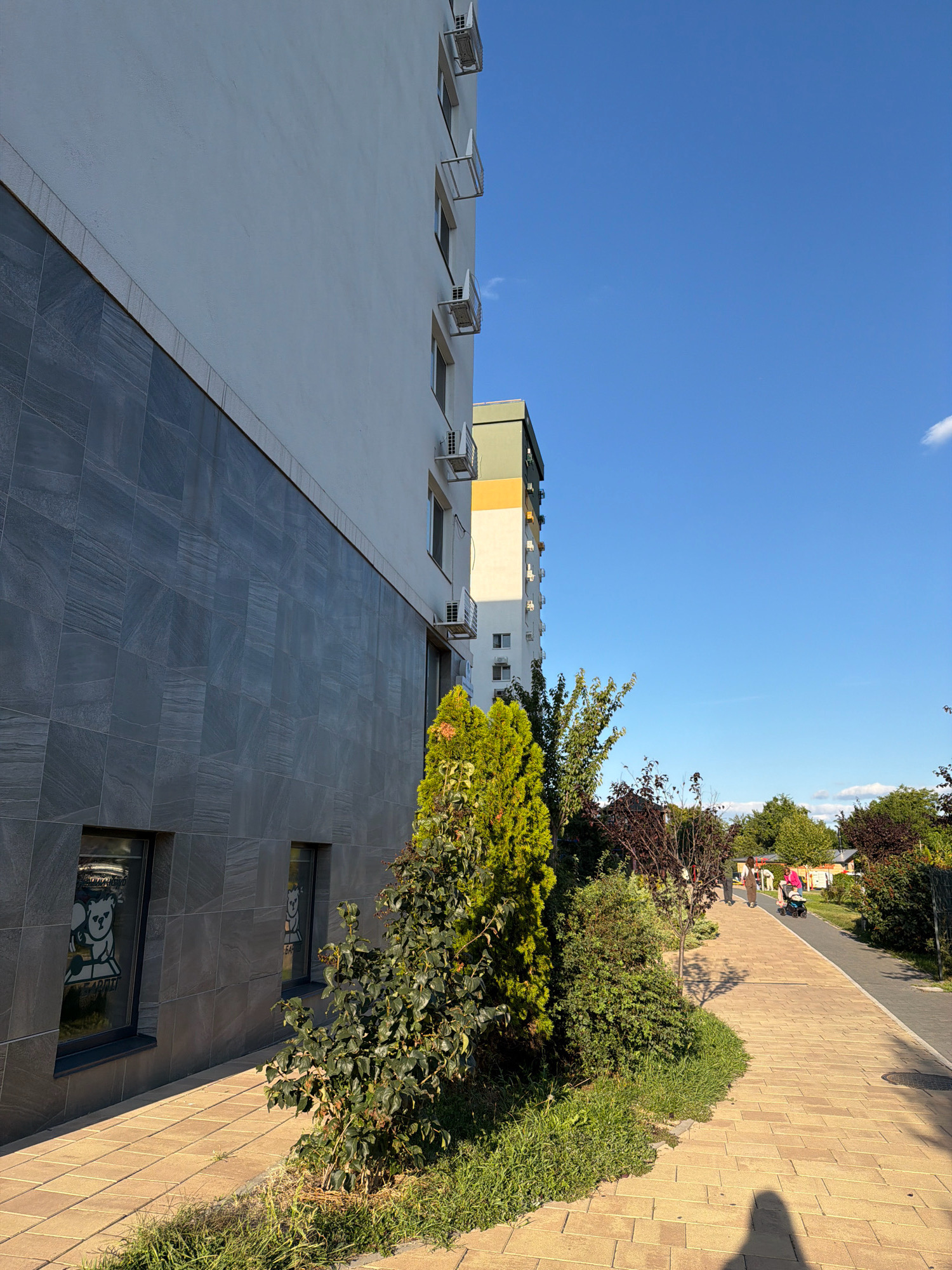 In Kyiv, Ukraine, on a sunny day, a long apartment building with a modern gray tiled facade stands to the left. The building has multiple air conditioning units visible on its exterior. A paved walkway runs alongside the building, bordered by greenery including bushes and trees. Further down the walkway, several individuals are visible. One person is pushing a stroller, and another individual appears to be walking towards them. The sky is clear blue, with a few wispy clouds. In the windows of the building, there are decals of cartoon bears. The overall scene depicts a residential area with a pedestrian pathway and landscaping.