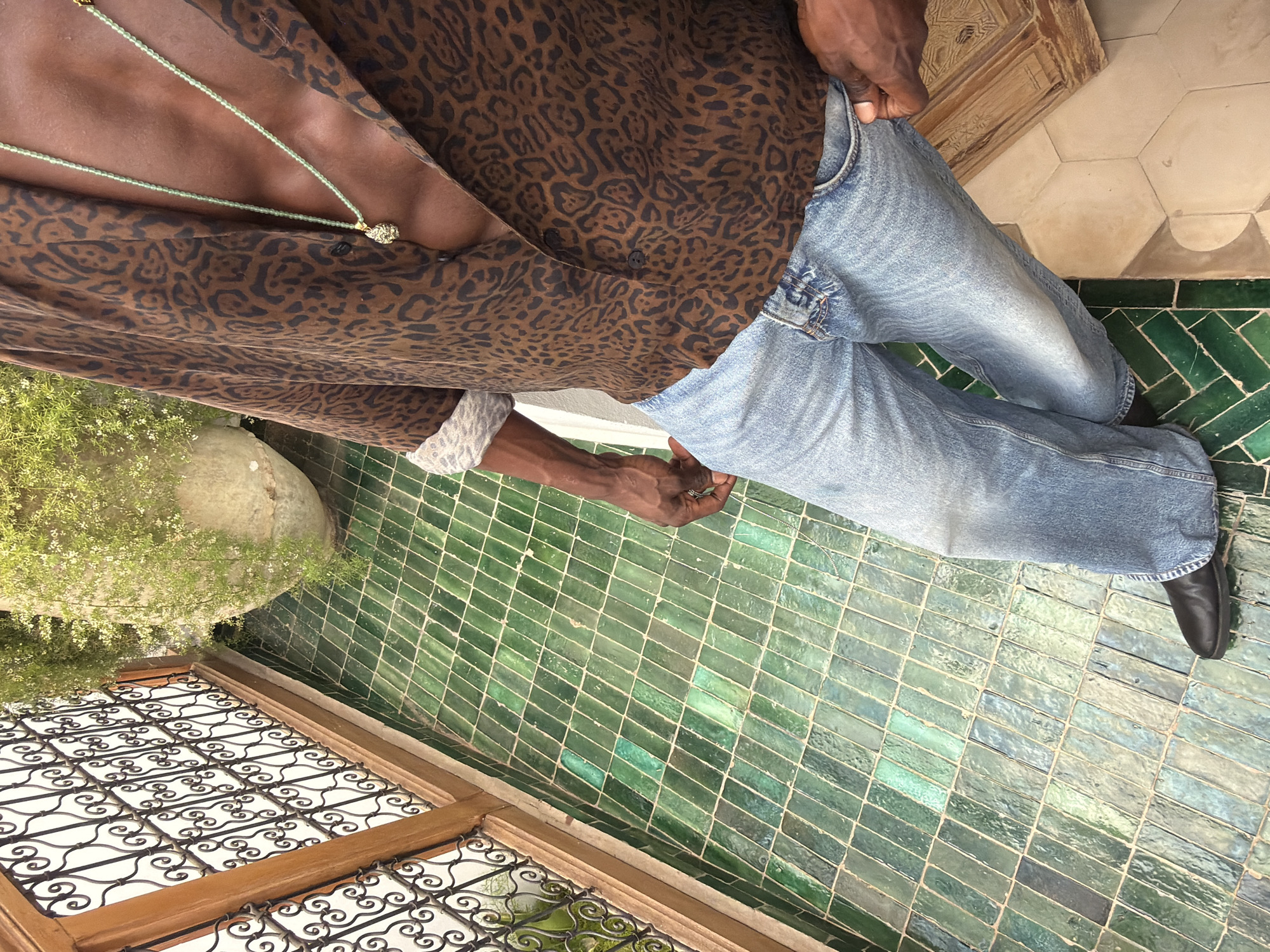 The image depicts a close-up, high-angle view of a person standing on a balcony in Marrakesh, Morocco. The balcony floor is covered in distinctive green zellige tiles, arranged in a herringbone pattern. To the left, a decorative wrought-iron railing with intricate scrollwork lines the edge of the balcony. Behind the railing, lush green plants spill from a large, weathered stone pot.
The person is wearing a brown, leopard-print short-sleeved shirt with the sleeves rolled up. A light green beaded necklace with a textured pendant is visible around their neck, resting against their chest. They are also wearing light-wash denim jeans. Their right hand is resting on their right hip, while their left hand is partially obscured by their jeans. Their left foot is clad in a black shoe. The overall aesthetic suggests a warm, outdoor environment, typical of a riad or traditional Moroccan courtyard.