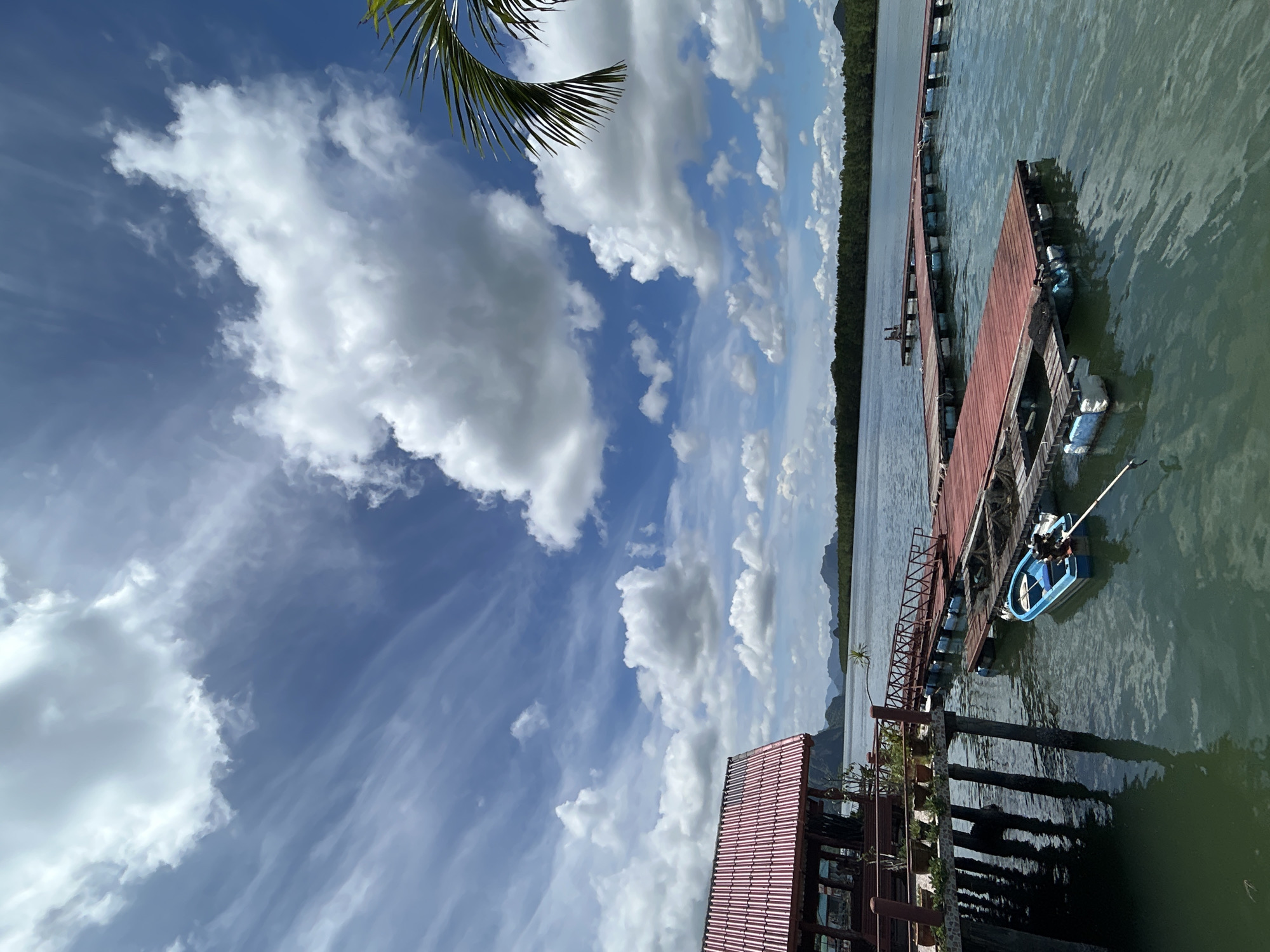 The image depicts a serene waterfront scene in Tambon Ko Panyi, Thailand, characterized by a bright, partly cloudy sky and calm, green water. In the foreground, a stilted building with a red corrugated roof and supporting posts stands on the left. Adjacent to it, a series of red wooden jetties extend into the water, some floating on blue buoys. A small, blue boat with an outboard motor is moored to one of the jetties by a white rope. The background features a dense line of green trees and distant, hazy mountains under a dynamic sky with wispy and puffy white clouds. A palm frond is visible in the upper right corner. The scene appears tranquil and suggests a location designed for leisure or fishing activities.