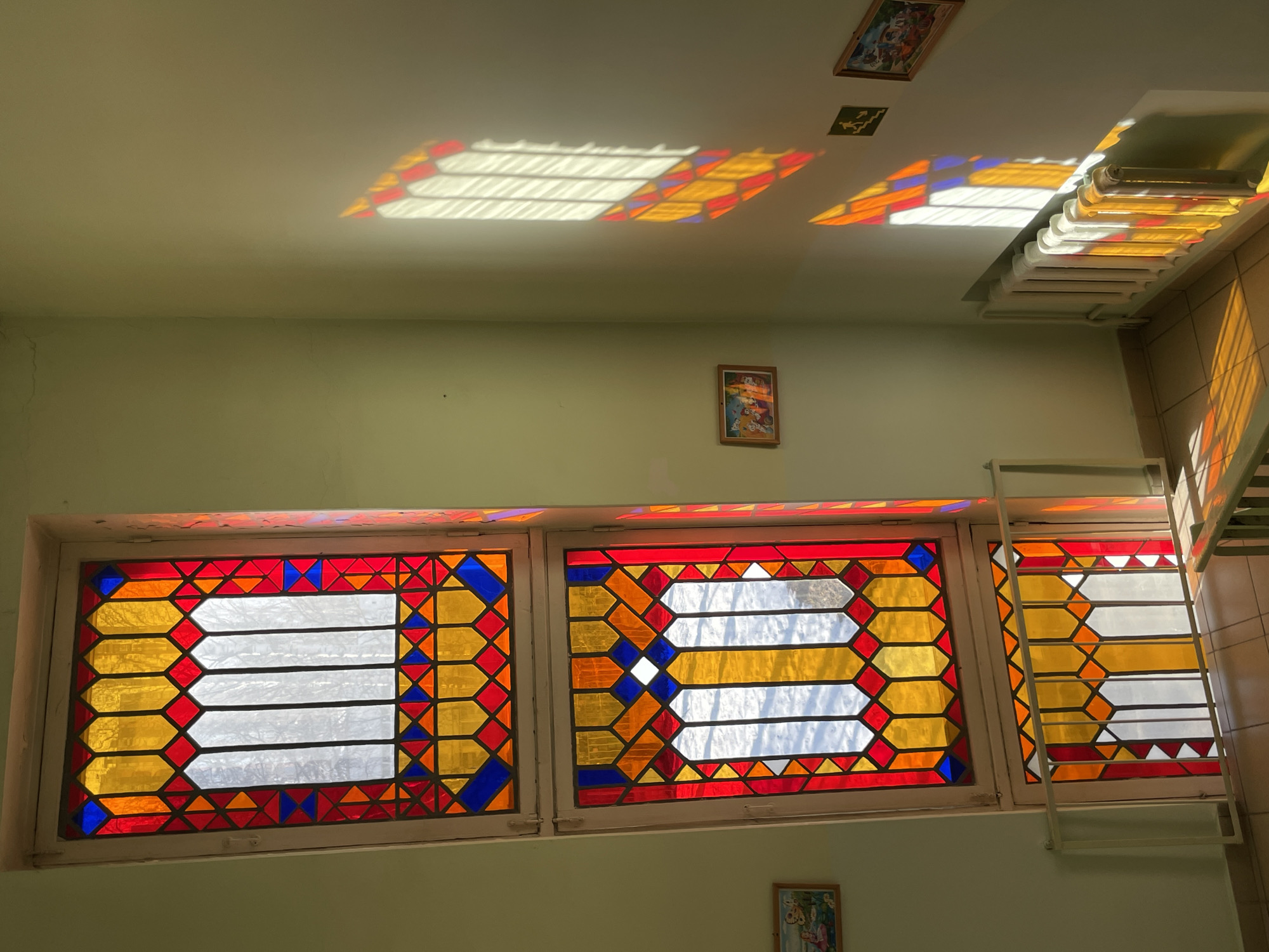 The image shows an interior view of a room in what appears to be a building in Izhevsk, Russia. The focal point is a set of three adjacent windows, each with a geometric stained-glass design featuring red, orange, yellow, and blue glass surrounding clear, horizontal panes. Sunlight shines through these windows, casting colorful geometric patterns on the ceiling. The walls are painted a pale green color. There are at least two small framed pictures hanging on the wall. Part of what seems to be a ladder is visible next to the right-hand window.