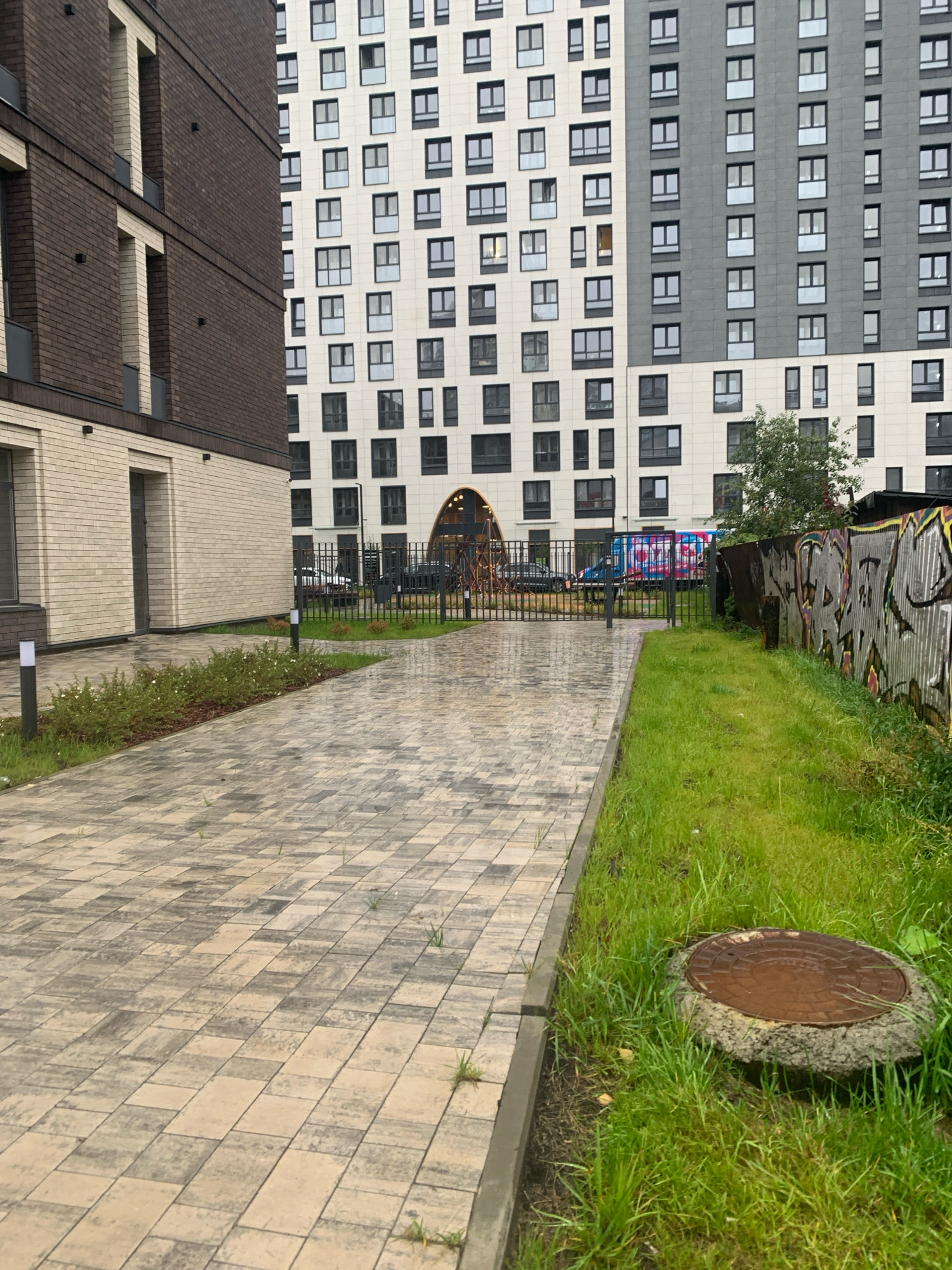 The image depicts a paved pathway in what appears to be a residential courtyard in Arkhangelsk, Russia. The pathway is made of interlocking rectangular pavers in shades of beige and gray, showing some signs of wear and with small weeds growing through the cracks. To the left of the pathway is a low-rise building with light-colored brickwork and dark-framed windows. Further in the background, a tall, modern apartment building with a grid-like facade of windows dominates the scene. To the right of the pathway is a patch of overgrown grass, a rusted manhole cover, and a corrugated metal fence covered in graffiti. A black metal fence separates the pathway from the background area where a vehicle and a truck with colorful decals are visible. The sky is overcast, suggesting a cloudy or rainy day, as evidenced by the wet appearance of the pathway. There are no people visible in the image. The environment appears to be a recently developed urban area.