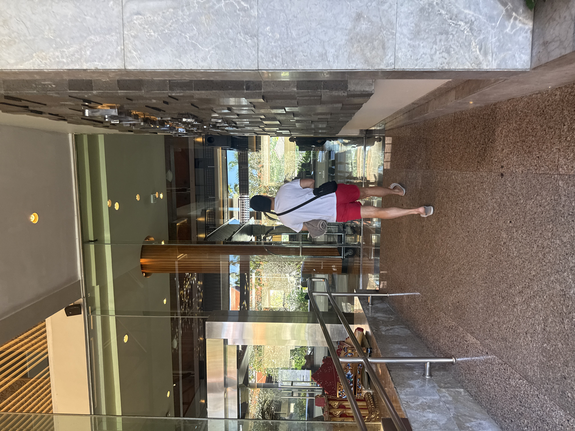 The image shows a man walking into the Westin Hotel in Kuta Selatan, Indonesia. He is wearing a white t-shirt, red shorts, and sandals. He has a black baseball cap and a shoulder bag. The hotel lobby is visible through the glass doors. The floor is a speckled brown material. The walls are decorated with stacked, rectangular stonework. There is a metal railing and some decorative items near the entrance.