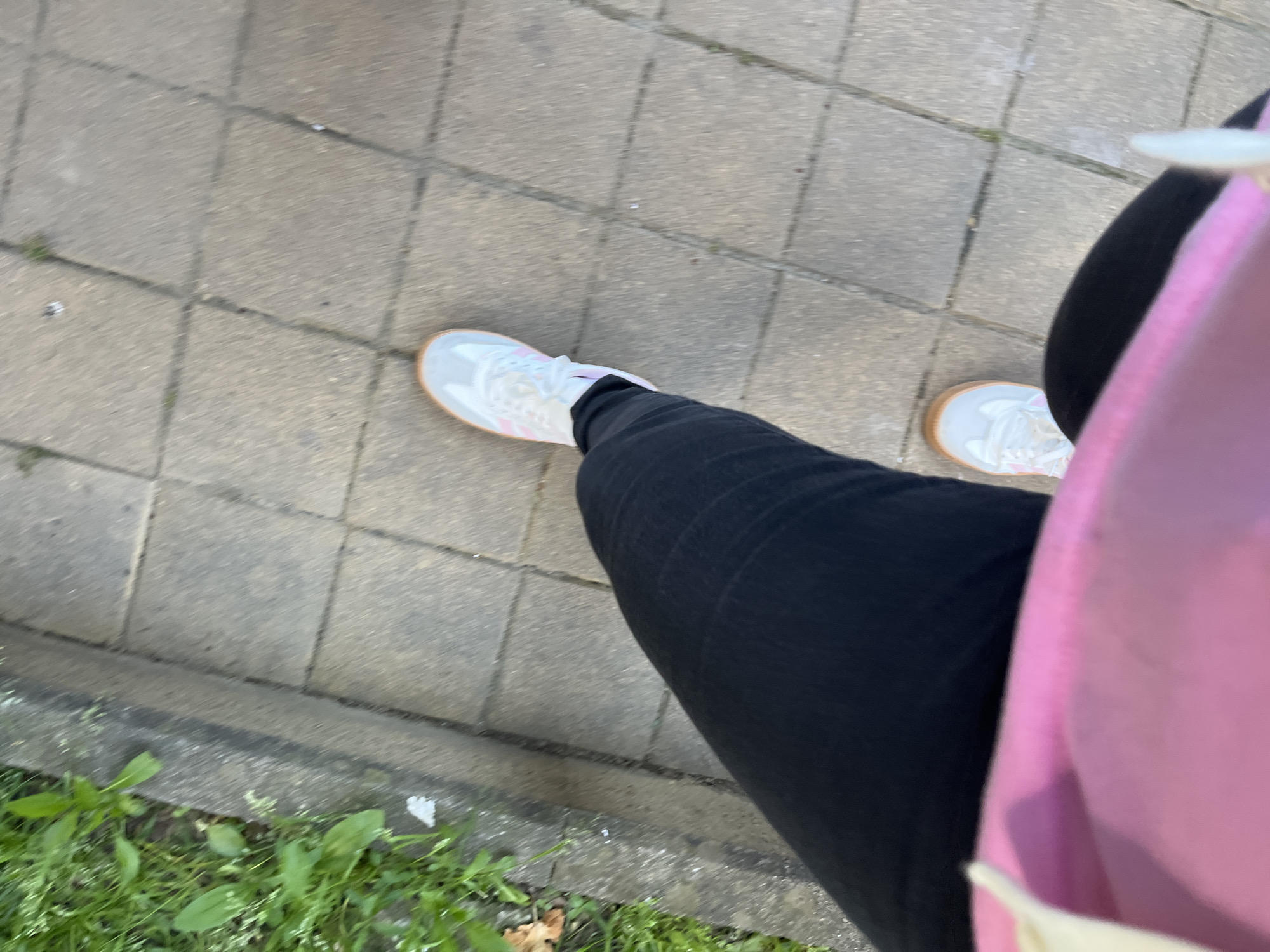 This first-person perspective image, taken in Sochi, Russia, shows the lower body of a person from an overhead view. The individual is wearing black pants and light-colored sneakers with pink details and tan-colored soles. A pink garment, possibly a top or a bag, is visible on the right side of the frame.
The setting appears to be an outdoor paved area, likely a sidewalk or pathway, constructed from grey rectangular pavers. Along the lower-left edge of the frame, there is a narrow strip of green grass and weeds, indicating the boundary of the paved surface, possibly next to a curb or green space. The lighting suggests it is daytime with clear, natural illumination. The image has a slight blur, which might indicate the person is in motion, walking along the path. No specific event or discernible emotion is captured, and there is no visible text.