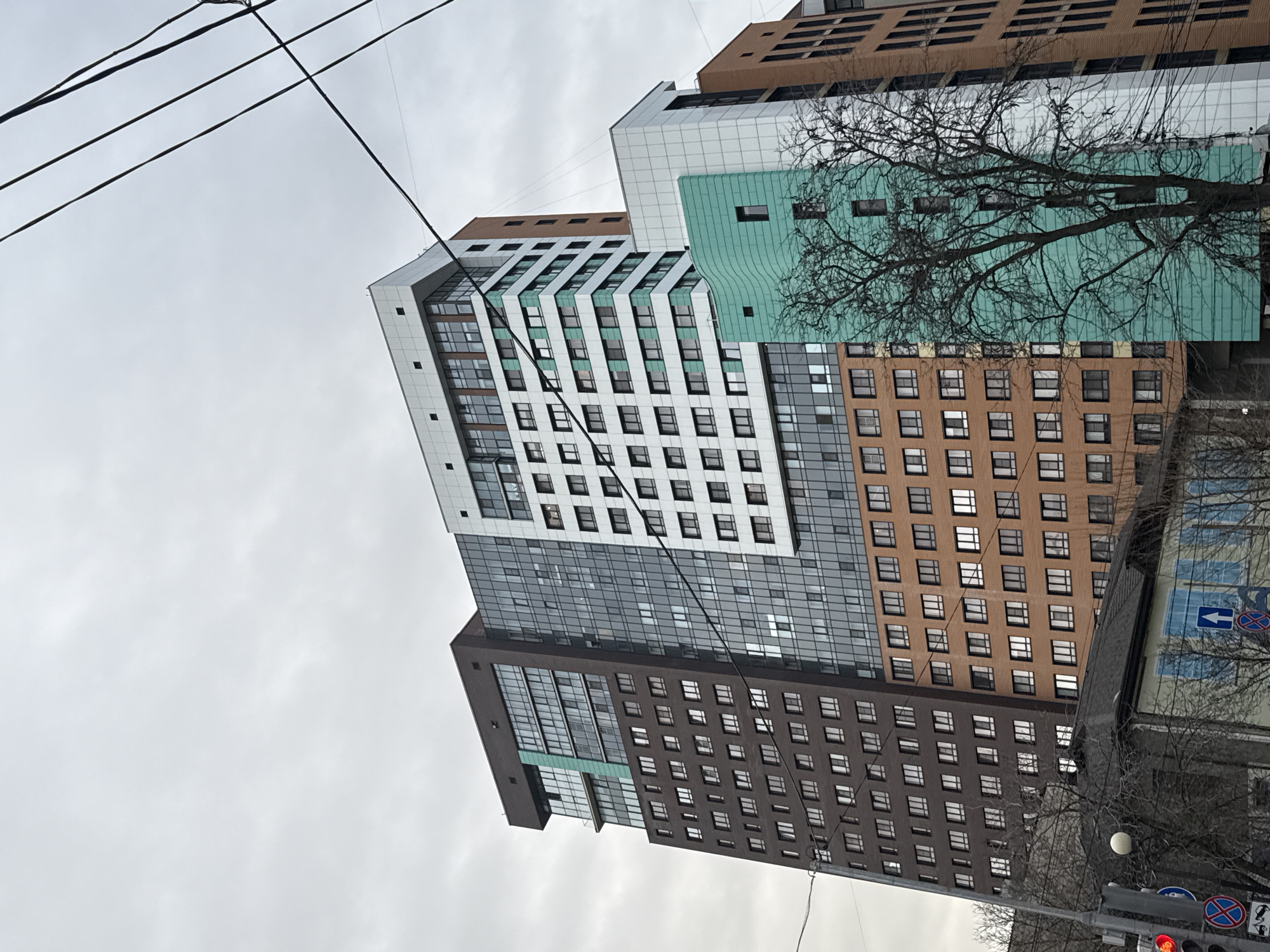 The image shows a cluster of modern buildings in Ovoshchnoy, Russia, viewed from a low angle, possibly street level. The buildings exhibit a mix of architectural styles and materials, including white, brown, and green paneling. The buildings are highrises or skyscrapers. Wires are visible in the sky, and a bare tree branch partially obscures one of the buildings. Traffic lights and street signs are visible at the bottom right. The sky is overcast. The buildings are arranged in a manner that suggests an urban setting.