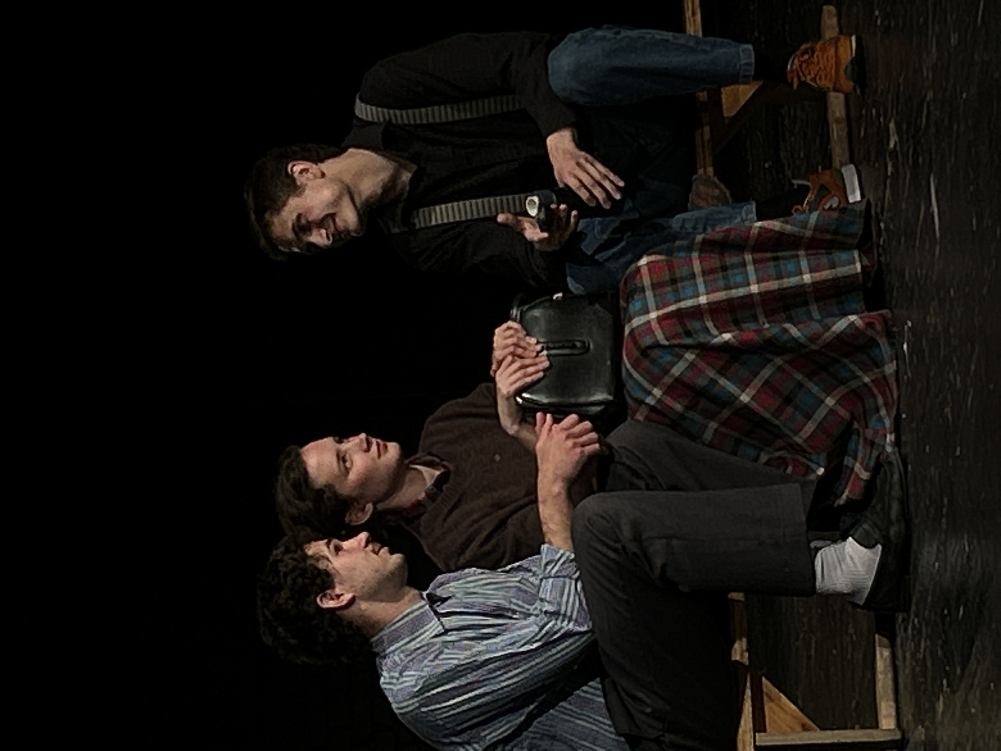 Three individuals are seated on a wooden bench against a black background. On the left, a male with dark curly hair wears a blue and white striped collared shirt and dark pants. He is looking to his right. In the center, a female with dark hair tied back wears a brown sweater and a plaid skirt. She is looking to her right and holding a black handbag. On the right, a male with dark hair wears a black collared shirt with suspenders and blue jeans. He is looking to his left and holding a small, dark object in his right hand. The lighting is focused on the individuals, with the background in shadow.