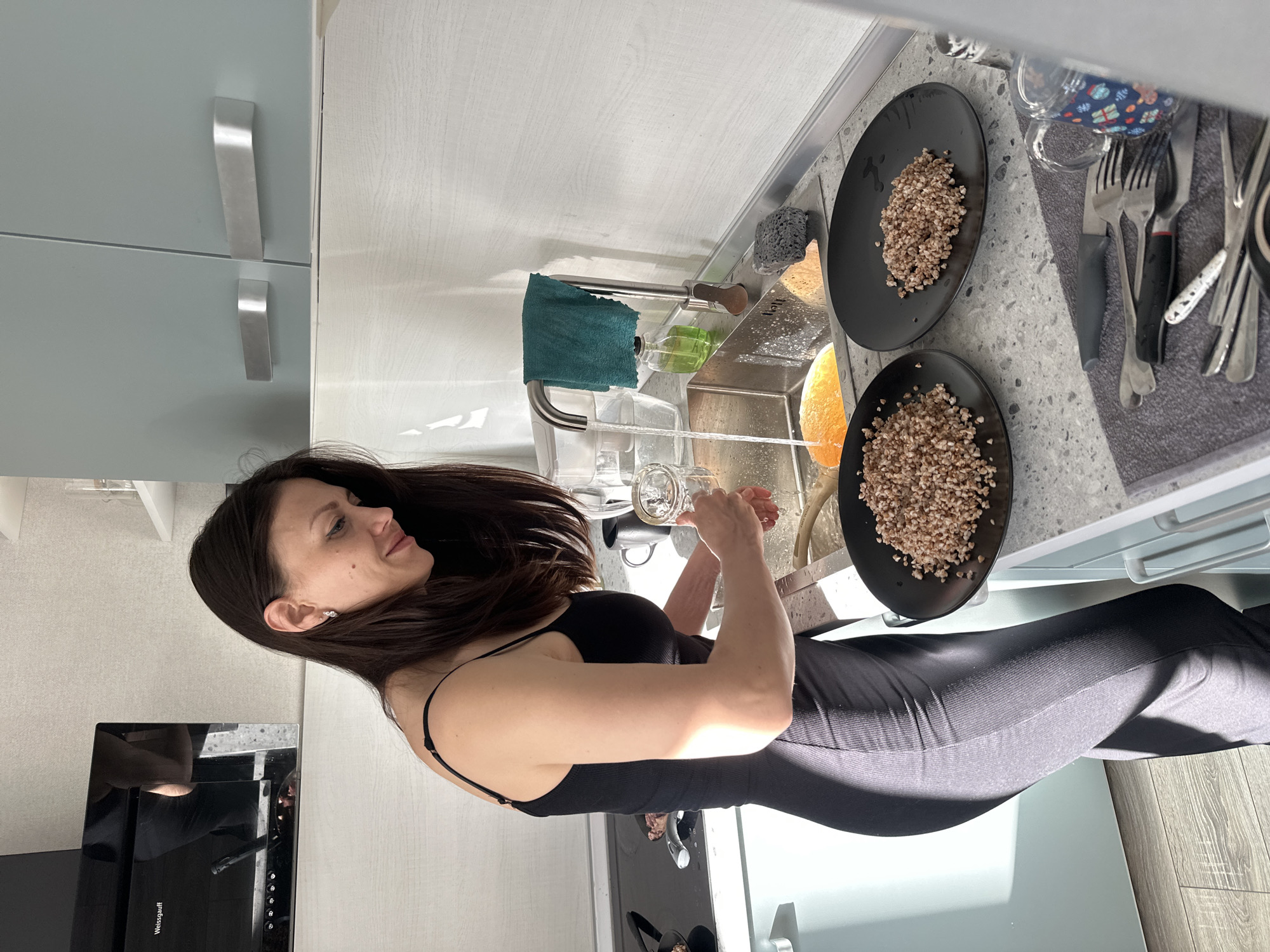 A woman is standing at a kitchen sink. She has long, dark hair and is wearing a black top and black pants. She is rinsing glasses in the sink. To her right, two black plates sit on the counter, each containing a portion of cooked grains. Behind the plates, there are assorted utensils including forks, knives and spoons, lying on a grey towel. The kitchen features light-colored cabinetry. A modern oven appliance is visible on the lower left corner. The scene appears to be set in a domestic kitchen. There is no verifiable evidence to confirm the location is Murino, Russia.