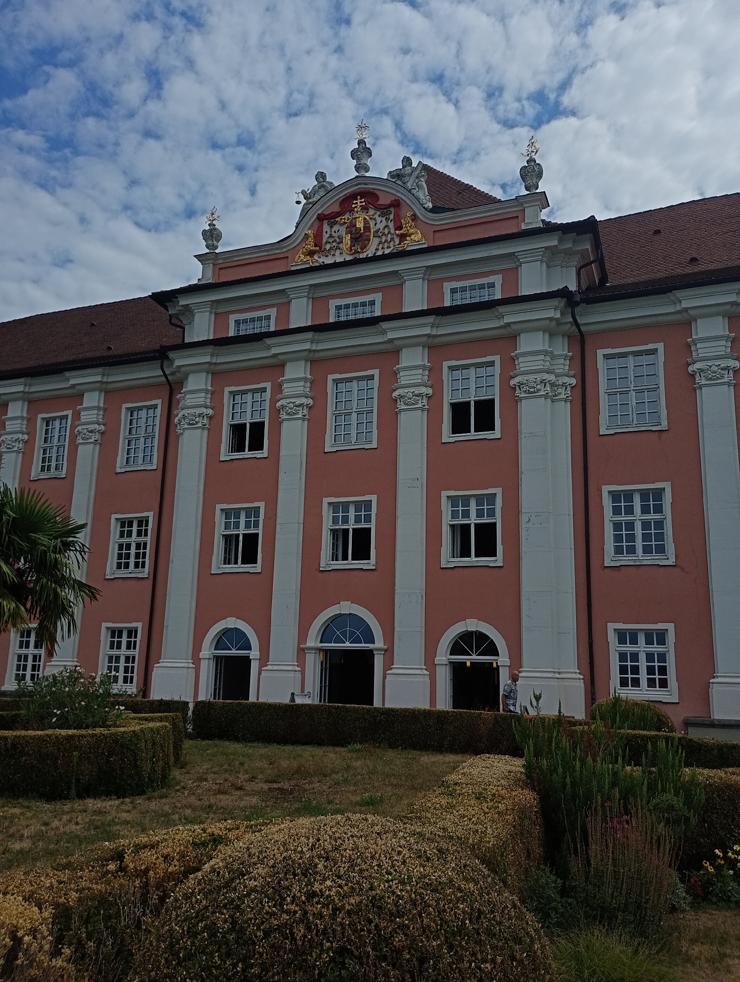 The image depicts the Neues Schloss, also known as the Prince-Bishop's New Palace, located in Meersburg, Germany. The building features a salmon-pink facade with white architectural details, including columns and window frames. The upper part of the building displays an ornate pediment with a coat of arms and decorative statues. Numerous windows are visible on different floors, some open and some closed. In the foreground, a meticulously landscaped garden with neatly trimmed hedges and various plants is present. A palm tree is visible on the left side of the image. A man in a plaid shirt is seen standing near an open doorway on the ground floor. The sky above is partly cloudy.