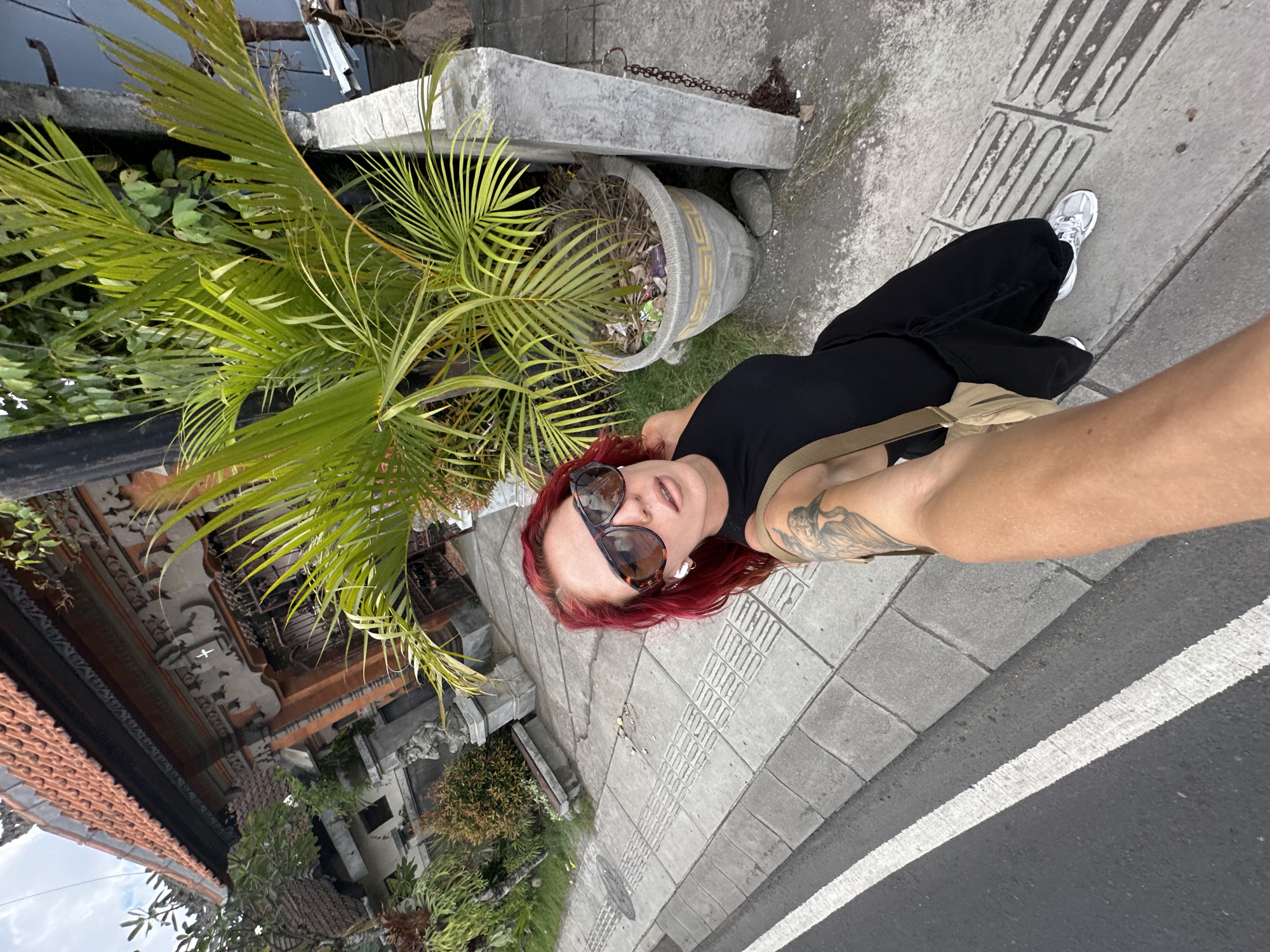 A woman with red hair and sunglasses is taking a selfie on a street in North Kuta, Indonesia. She is wearing a black top and pants with white sneakers. Her left arm is extended towards the camera. On her right arm, a tattoo is visible. Beside her is a large potted plant and behind her is a building of traditional Balinese architecture. The ground is paved with grey concrete tiles. The selfie is taken with the camera facing down.