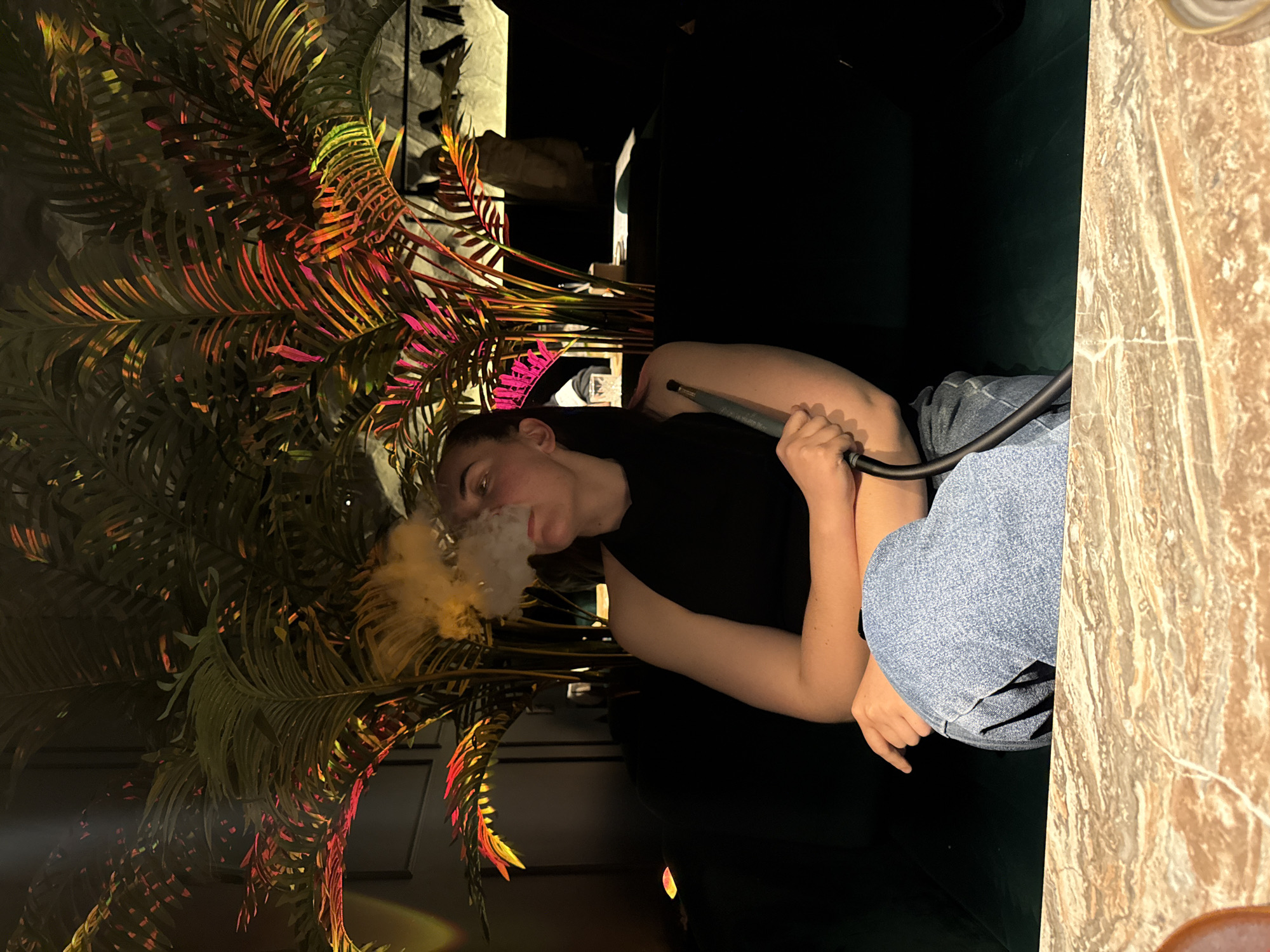 A woman is smoking a hookah in a dimly lit interior. The woman, wearing a black sleeveless top and denim jeans, exhales smoke. She holds the hookah hose in her right hand. Decorative leafy plants surround her, illuminated with colorful lights. The edge of a marble table is visible on the right side of the image. The location is possibly Chaikovsky, Russia, based on the prompt.