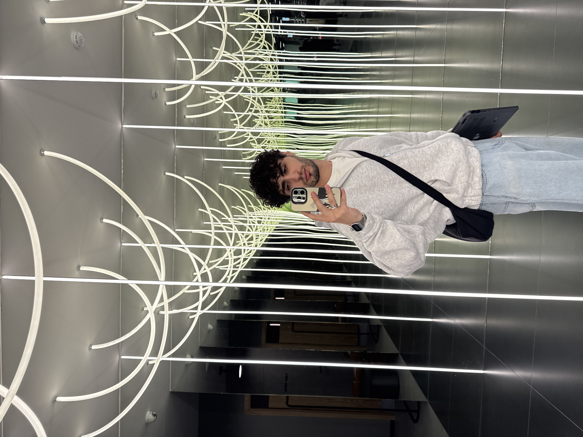 A young man is standing in an elevator with mirrored walls, taking a selfie with his smartphone. He has curly dark hair and a light beard. He's wearing a light grey sweatshirt, light blue jeans, and a black shoulder bag. He's holding a black tablet in his other hand.
The elevator's walls are lined with horizontal rows of white neon lights. Curved neon lights adorn the side walls. The floor is dark, and the reflection in the mirrors creates an illusion of a never-ending corridor.
The elevator appears to be located in a modern building in Yerevan, Armenia.