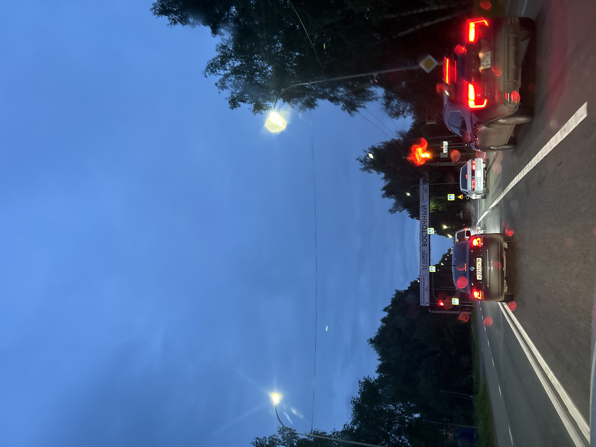 The image depicts a twilight scene on a multi-lane asphalt road in Italmas, Russia, viewed through a vehicle's windshield speckled with water droplets, indicating recent or ongoing precipitation. The sky is a deep blue, with scattered clouds. Streetlights on both sides of the road are illuminated, casting yellow-white light and exhibiting lens flare due to the wet windshield.
Overhead, a gantry structure spans the road, displaying Russian text signs. The prominent signs read "ФИРМЕННЫЙ МАГАЗИН" (Company Store), "КОМОС" (KOMOS), and "ВОСТОЧНЫЙ" (Vostochnyy/Eastern). Below these, a "СТОП" (STOP) sign is visible. Red traffic signals are also illuminated on the gantry, indicating a stop. Pedestrian crossing signs (blue squares with white figures) and yellow diamond-shaped cautionary signs are present along the road.
The road features clear double white lines separating the lanes. Dense, dark green trees line both sides of the road, receding into the distance.
In the left lane, a dark-colored Volkswagen Polo sedan is stationary, displaying brightly illuminated red brake lights. Its rear license plate, "x331cy 18," is clearly visible, with "18" corresponding to the Udmurt Republic, where Italmas is located. Further ahead in the left lane, a white sedan, possibly a Toyota, is visible with its taillights on.
In the right lane, a dark red SUV is stationary, with its bright red brake lights also illuminated. Its rear license plate is partially obscured and difficult to read definitively, but visible characters include "x74". A white van or large SUV is partially visible behind it.
No individuals are discernible outside of the vehicles. The scene captures an evening traffic stop on a wet road, characterized by artificial light sources contrasting with the dim natural light.