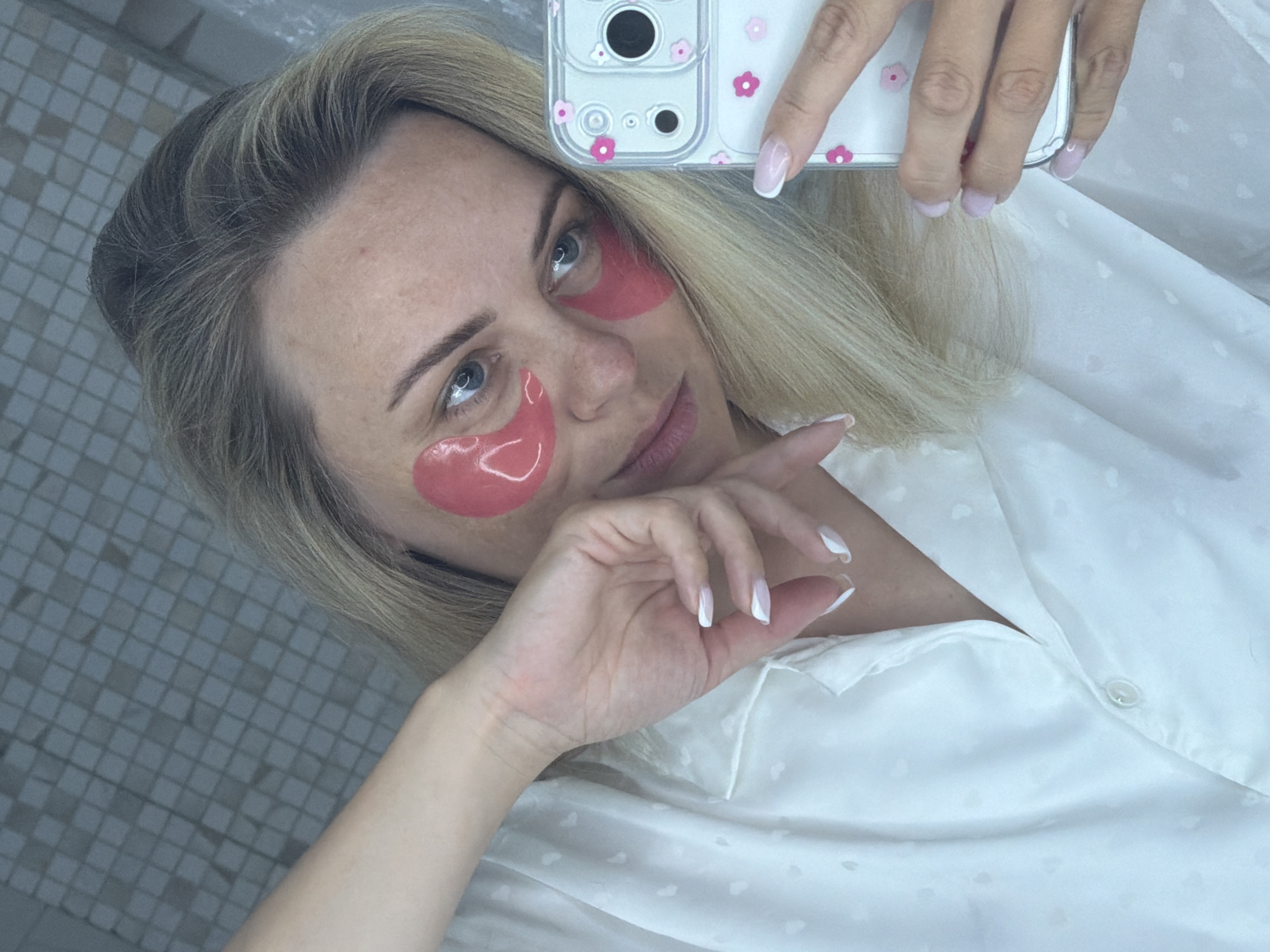 A woman is captured in a selfie, likely in an indoor setting such as a bathroom or bedroom in Votkinsk, Russia, given the tiled background. She holds a smartphone with a clear case decorated with pink flowers, positioning it to take the picture. Her blonde hair is loosely styled around her face, and she gazes towards the camera with a soft, relaxed expression and a faint smile.
She is wearing pink, hydrogel under-eye patches, suggesting a skincare routine or a moment of self-care. Her hands are visible, showing manicured nails with a white French tip design. She wears a white, collared shirt made of what appears to be a silky or satin fabric, featuring a subtle, embossed pattern of small hearts or dots. The background consists of light-colored, square tiles. The overall scene conveys a personal and tranquil moment.