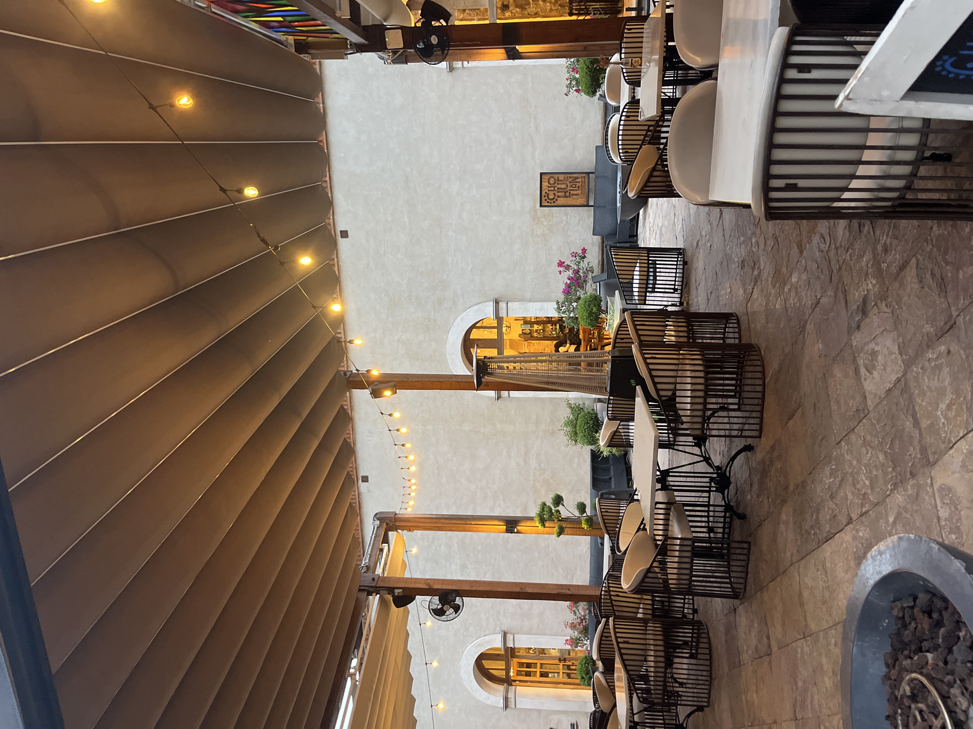 This is an outdoor courtyard or patio of a business in what seems to be Domingo Arenas, Mexico, based on architectural cues. The area is covered by a retractable awning strung with decorative lights. The space is furnished as an outdoor dining area, with tables and chairs arranged in rows. The chairs are a distinctive design, with metal cages forming the base and white curved seats. A sign with the name of the establishment is visible on the wall. The building features arched windows and doors, indicating a historical or traditional style. The ground is paved with stone. A fire pit is situated to the side, filled with dark stones or lava rocks. There are no people visible in the scene, and the space appears empty.