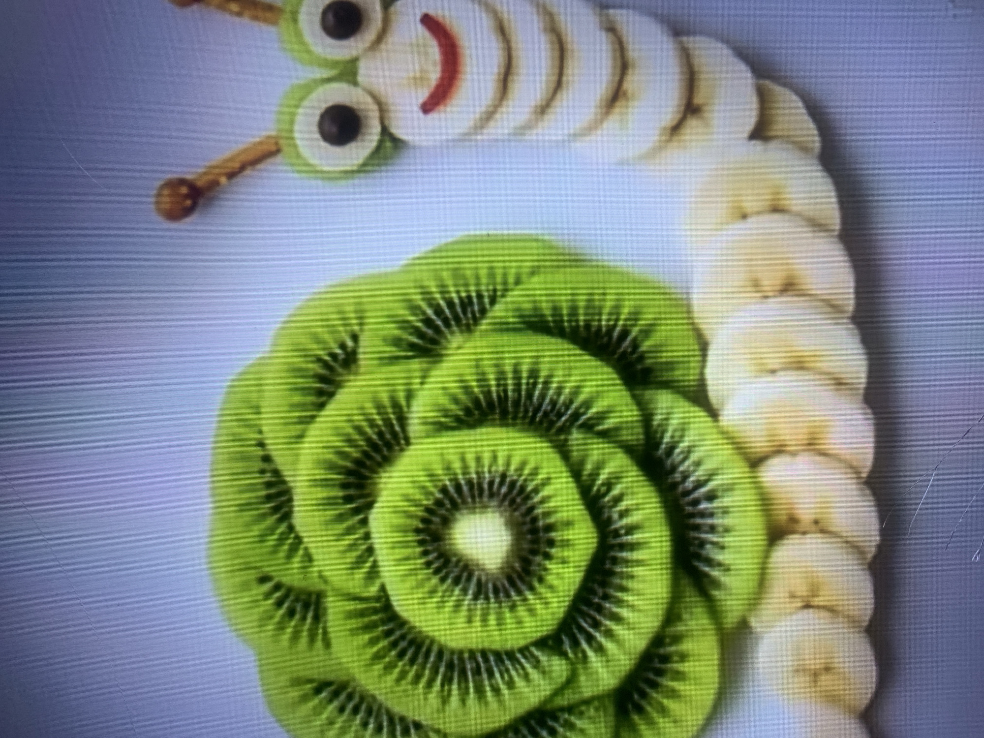 The image displays a close-up, overhead view of a fruit-shaped snail. The snail's body is composed of sliced bananas arranged in a segmented, linear pattern, curving towards the right. The snail's head features two large, green circular pieces of kiwi fruit acting as eyes, with dark brown circular pieces of fruit or candy for pupils. A curved, red line resembling a mouth is positioned below the eyes. Two pretzel sticks with a brown, rounded end are placed above the eyes, representing antennae. The snail's shell is intricately constructed from numerous overlapping slices of kiwi fruit, arranged in a spiral pattern, forming a large, circular shape on the left side of the image. The background is a plain, light blue or gray surface. There is no discernible indication of the city of Almaty, Kazakhstan, or any urban environment.