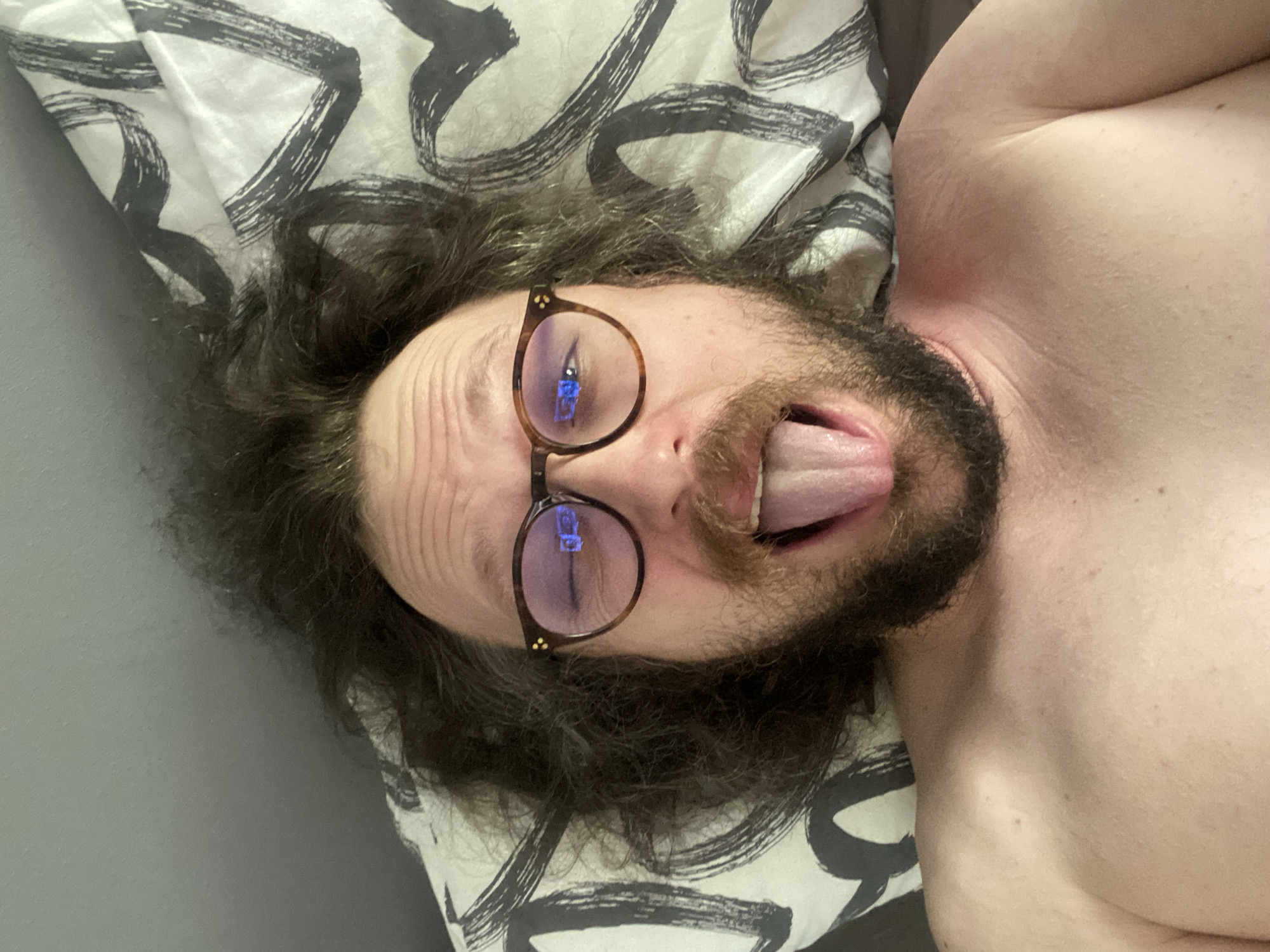 A selfie of a man with long, dark, messy hair, a beard, and a mustache is taken from a low angle. He is lying down, with his head on a pillow that has a black and white abstract design. He is wearing eyeglasses with tortoiseshell frames and his mouth is open, with his tongue sticking out. His left eye is partially closed, and his right eye is visible through his glasses, reflecting a blue light. His bare chest and shoulders are visible in the lower part of the frame. The background consists of a gray surface to the left and the patterned pillow to the right. The image was taken in Rusanovo, Russia.