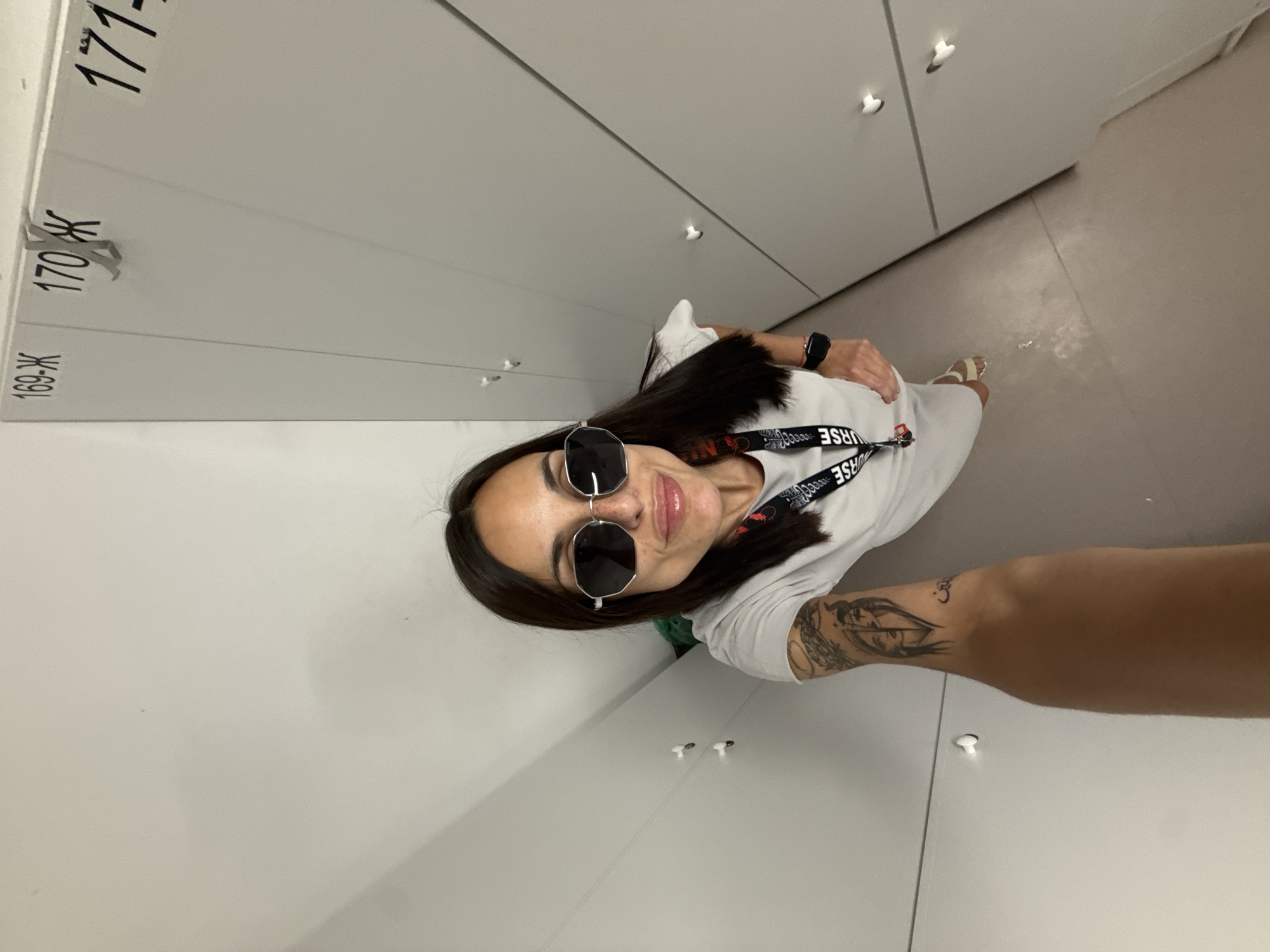 The image is an indoor selfie taken in what appears to be a locker room. The person taking the selfie is a woman with dark hair and wearing sunglasses. She has a white t-shirt on and a lanyard around her neck that has the word "NURSE" printed on it. She also has a tattoo on her left forearm.
The background consists of white lockers with numbers on them: "169-Ж", "170 Ж", and "171". The room has a tile floor.
Without further information, I am unable to confirm the location in the city of Pleven, Bulgaria.