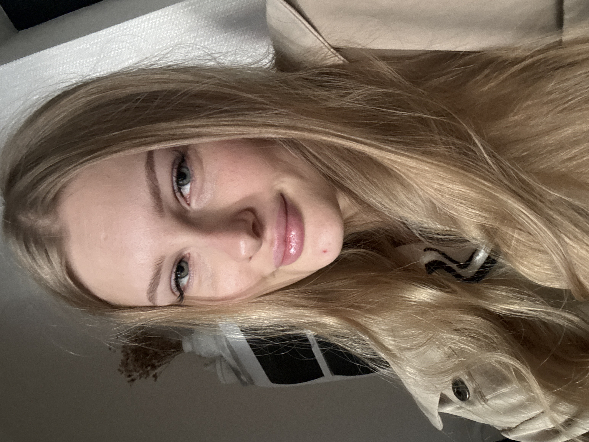 The image is a close-up, selfie-style photograph of a young Caucasian woman with long blonde hair. She is wearing a light-colored trench coat. Her facial features are clearly visible, including blue-green eyes, full lips with pink lipstick, and fair skin. There is a small red mark on her chin. The background is softly blurred, showing a hint of a white curtain or blinds on the right and a dark object and some dried flowers in a vase on the left. The lighting suggests an indoor environment, possibly a room. There is no information in the image to determine its location in Perm, Russia.