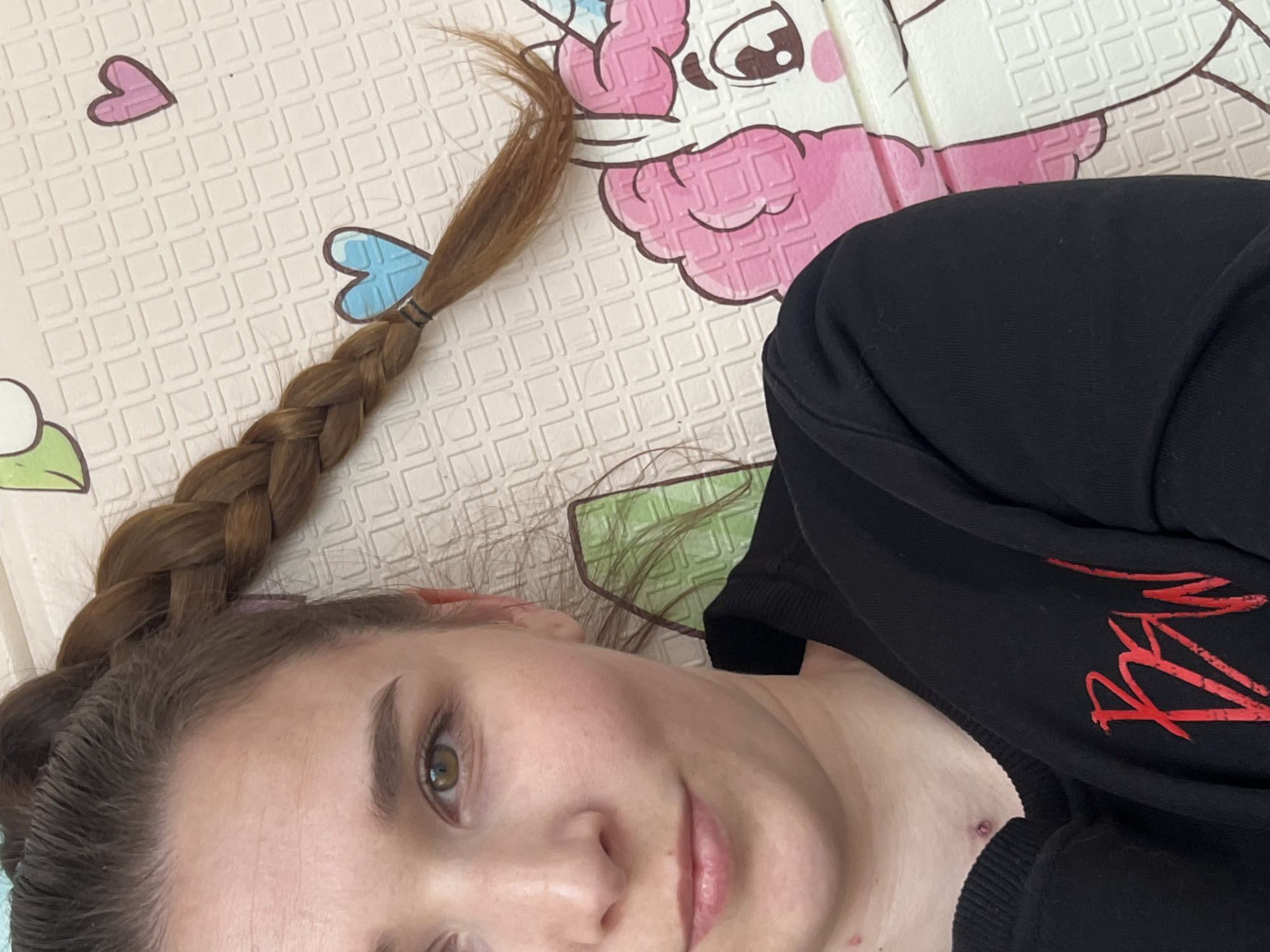 The image shows a young woman lying down, looking towards the camera with a calm, slightly smiling expression. Her head is angled, and her gaze is directed upwards. She has fair skin, light brown eyes, and dark eyebrows. Her long, light brown hair is braided into a single plait, which rests on the mat beside her.
She is wearing a black long-sleeved top or sweatshirt. On the front of her top, there is a design of stylized red text or graphics, which is partially obscured and not fully legible. It appears to be composed of angular, somewhat jagged lines. Minor blemishes are visible on her neck and chest area.
The background consists of a light-colored, padded mat with cartoon patterns. Visible elements of the pattern include small pink and blue hearts, and parts of larger cartoon characters such as a pink, large-eyed creature resembling a unicorn or pony, and a green character (possibly a frog) on the lower left. The mat itself has a subtle raised grid texture. The setting appears to be indoors, likely in Votkinsk, Russia, and the lighting suggests it is daytime. The overall scene conveys a casual and relaxed atmosphere.