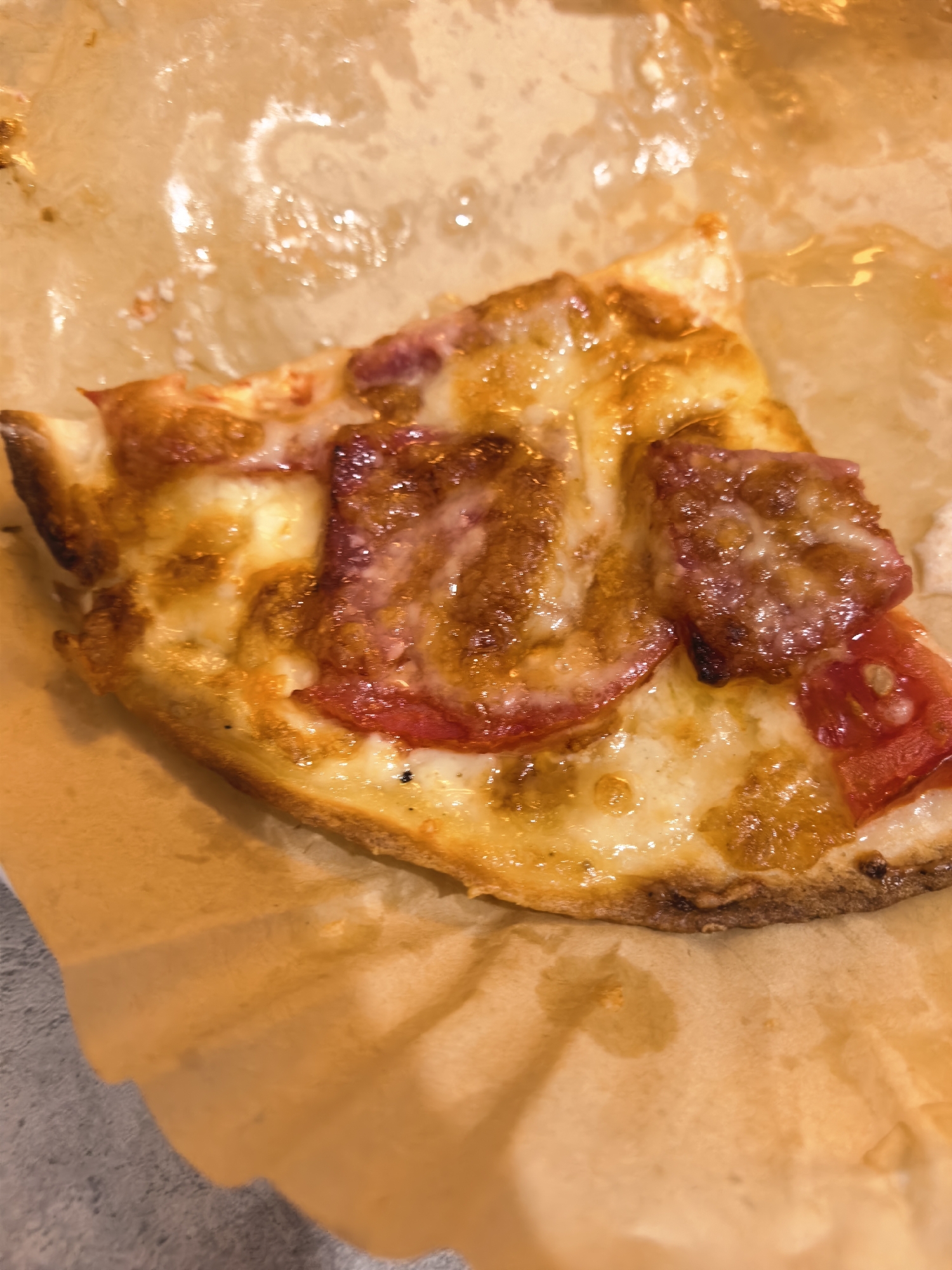 A close-up shot shows a slice of pizza on brown paper. The pizza has a golden-brown crust with melted cheese and red toppings. The paper is crinkled and shows grease stains. The background is blurred, but appears to be a countertop.
