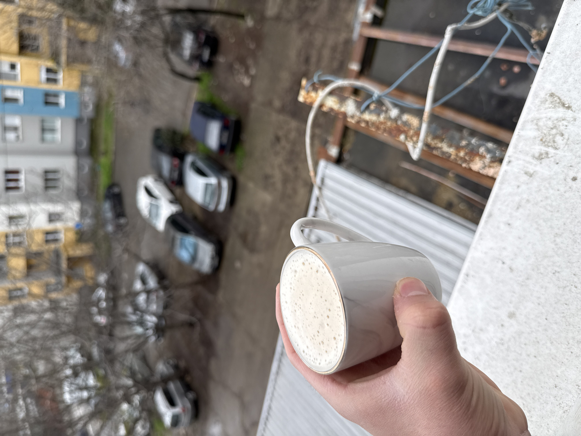 The image shows a first-person perspective of a hand holding a white and gold rimmed mug filled with a light-colored beverage, likely a cappuccino or latte. The backdrop reveals an elevated view of an urban environment.
From the vantage point, one can see the rooftops of adjacent buildings and parked vehicles. A nearby balcony railing and clothes drying rack are visible. In the background, blurred building facades and trees suggest a residential area.
Based on research of the area, the building with colored walls (yellow, blue, and gray) is located at ул. „Генерал Столетов“ 93, 6000 Stara Zagora Center, Stara Zagora, Bulgaria.