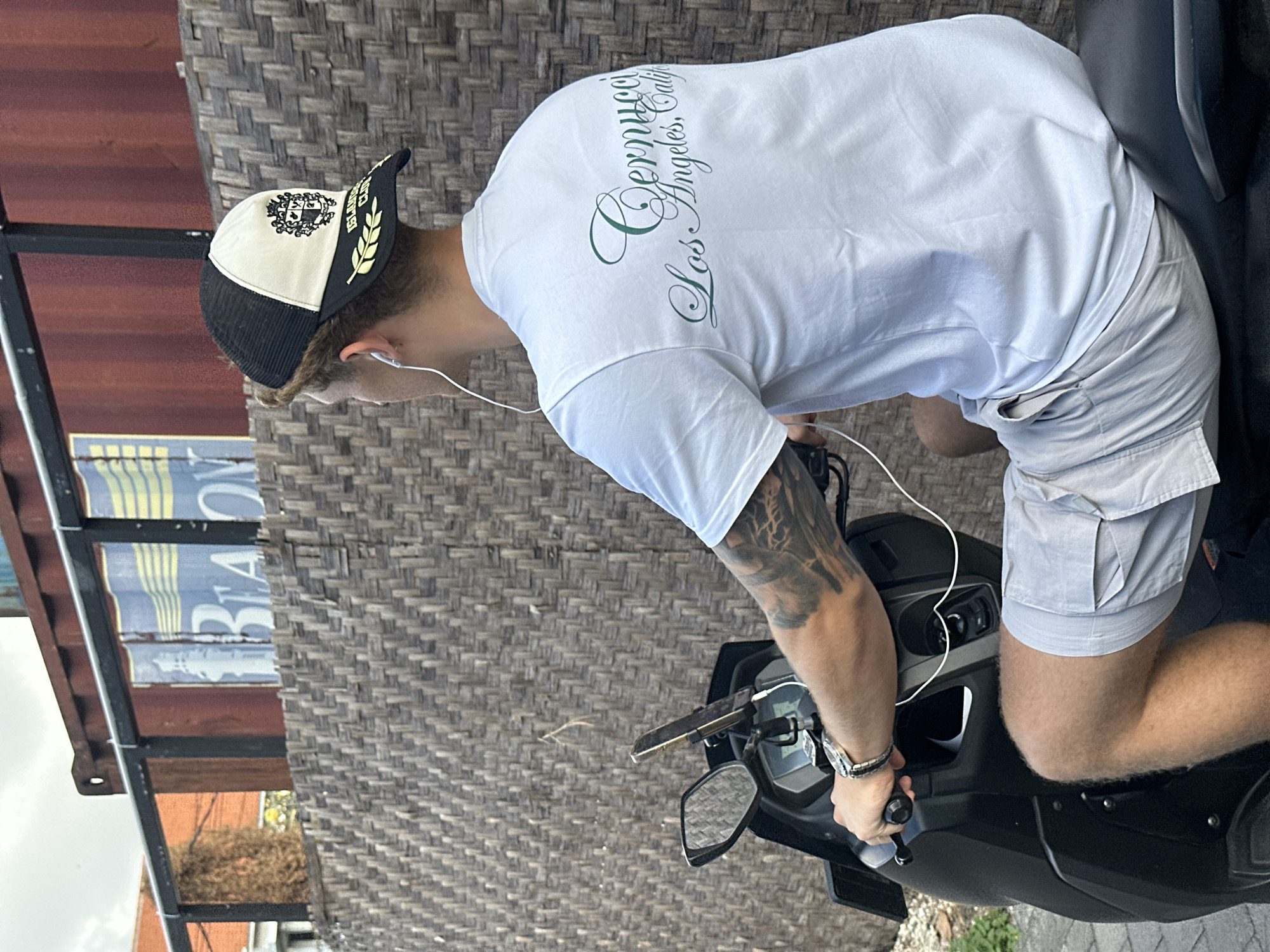 A young man with visible arm tattoos is riding a dark-colored scooter. He is wearing a white t-shirt with green script text that reads "Cernucci Los Angeles, Calif" and light gray cargo shorts. On his head, he wears a black and beige baseball cap with a crest and laurel leaf design. He is also wearing white earbuds. The scooter has a phone mount on the handlebars. Behind the man, there is a textured woven wall made of light brown reeds. Beyond the wall, a weathered shipping container with a black metal railing is visible, displaying a faded blue and yellow sign with white lettering. The background also shows part of an orange building with a dense, dry bush. The scene appears to be outdoors, possibly in a street or courtyard environment in North Kuta, Indonesia.