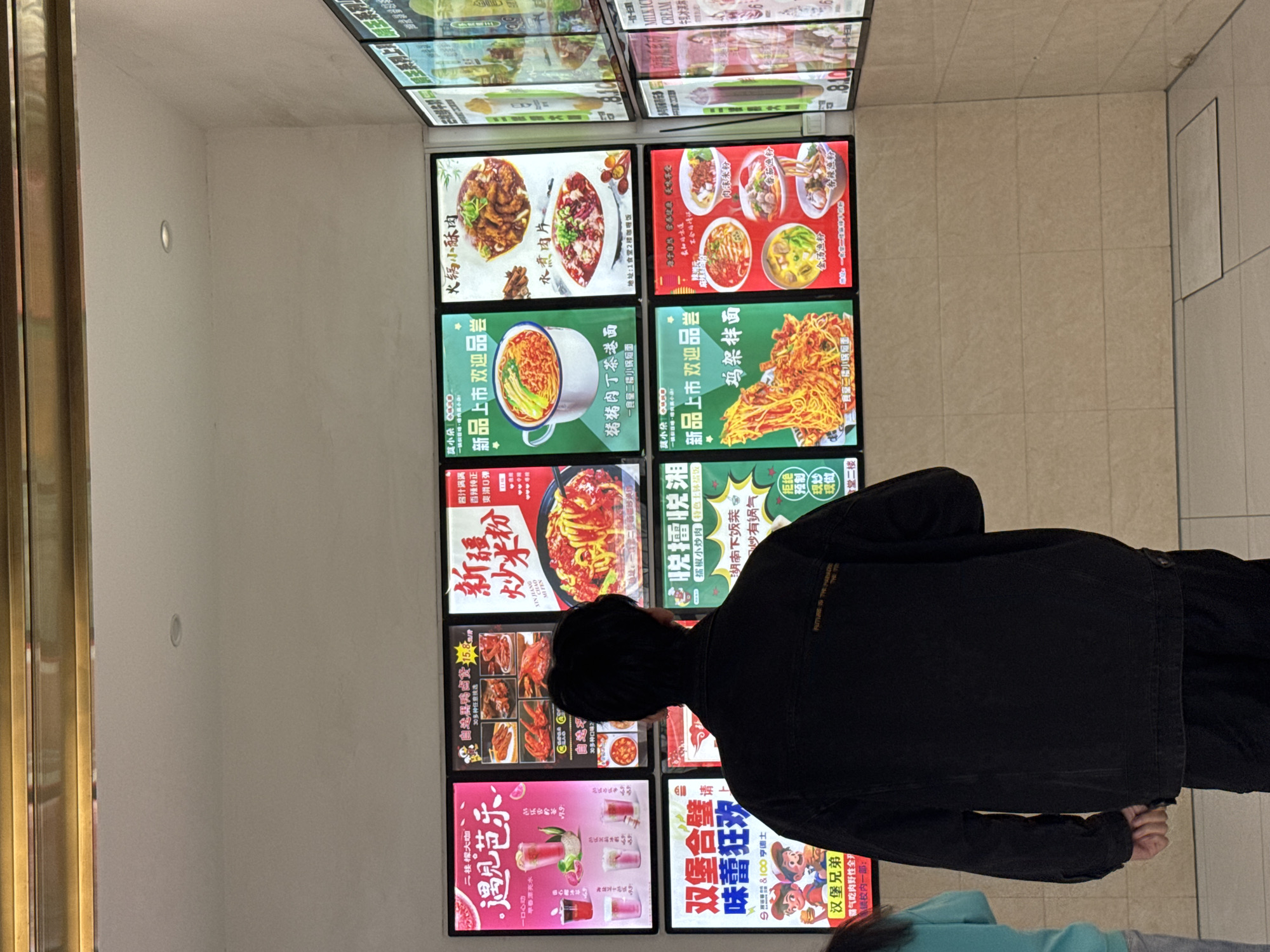 Here's a detailed description of the image:
**Scene Overview:**
The image shows a person standing in front of a wall covered with illuminated food advertisements. The scene appears to be indoors, likely in a food court or restaurant area.
**Location:**
Based on the text in the ads, the location is likely in China. Zhongtun, China is not identifiable, so the location of this photo cannot be determined beyond China.
**People:**
* One individual is prominently featured, wearing a black jacket. This person seems to be looking at the advertisements. The person has black hair.
**Subjects/Objects:**
* **Advertisements:** A grid of illuminated food advertisements dominates the scene. The ads depict various dishes, including hot pot, noodles, braised meat, drinks, and other Chinese-style dishes. The ads contain Chinese characters and images of the food items.
* **Walls:** The walls are painted a light color, providing a neutral backdrop for the advertisements.
* **Other Objects:** The image includes a part of a door frame on the left side and, in the bottom right, glimpses of another person wearing a blue top and gray pants.
**Actions/Interactions:**
* The primary action is the person in the black jacket looking at the food advertisements, presumably deciding what to order or simply viewing the selections.
**Environment:**
* **Lighting:** The scene is well-lit, primarily due to the bright illumination of the advertisements.
* **Atmosphere:** The atmosphere suggests a casual dining environment.
