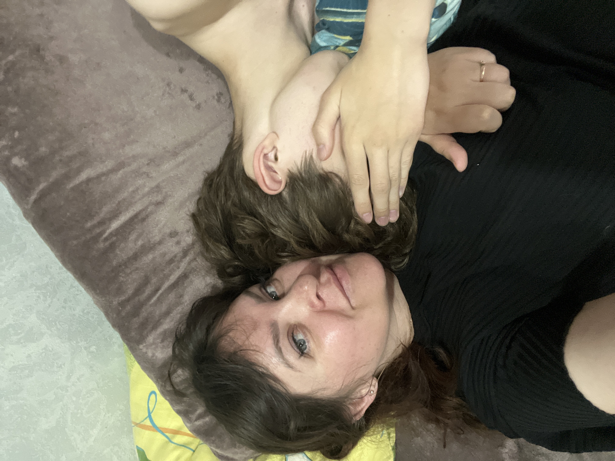 A photo depicts two people lying on a bed or couch. One person, presumably a woman, faces the camera and is lying on her back, with her head resting on a yellow and green patterned pillow. She has long, wavy dark hair and blue eyes. She wears a black top. The other person is partially obscured, with their head resting near the woman's shoulder and one of her hands covering their face. This person has short brown hair and is wearing a blue and green patterned shirt. The background consists of a brown textured surface, which may be a blanket or the couch itself. A ring is visible on the woman's finger. The location and city of Noviy, Russia cannot be ascertained from the image.