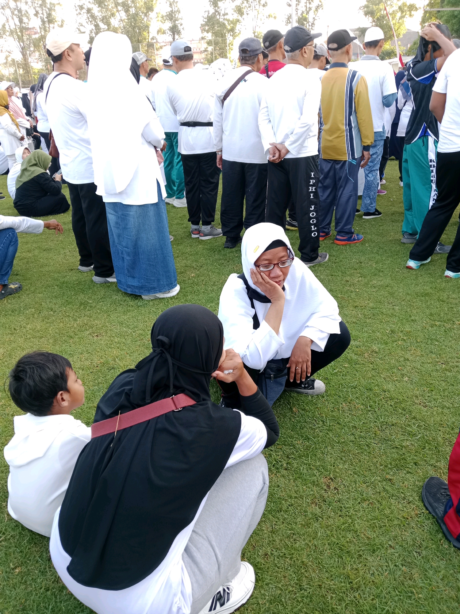 The image depicts an outdoor gathering on a grassy field, likely in Surakarta, Indonesia, as suggested by the text on some participants' clothing. The scene is populated by a diverse group of individuals, many wearing casual sportswear, indicative of a community event or recreational activity.
In the foreground, three individuals are the primary focus. A young boy in a white hooded shirt is looking to his left. Beside him, a woman wearing a black hijab and a white undershirt is seated, her back partially to the camera, with a red strap across her chest. Another woman, wearing a white hijab and glasses, is also seated, leaning forward with her hand on her cheek, appearing contemplative. She is dressed in a white top and black pants.
In the background, a larger crowd is visible, many standing. Several individuals are wearing white t-shirts and caps, suggesting a uniform for the event. Some are in athletic wear, including track pants with text that appears to be in Indonesian, such as "IPHI JOGLO" visible on one person's leg. The environment is characterized by a well-maintained grassy area with trees and foliage in the distance, suggesting a park or a large open space within the city. The lighting suggests it is daytime, with sunlight filtering through the trees.