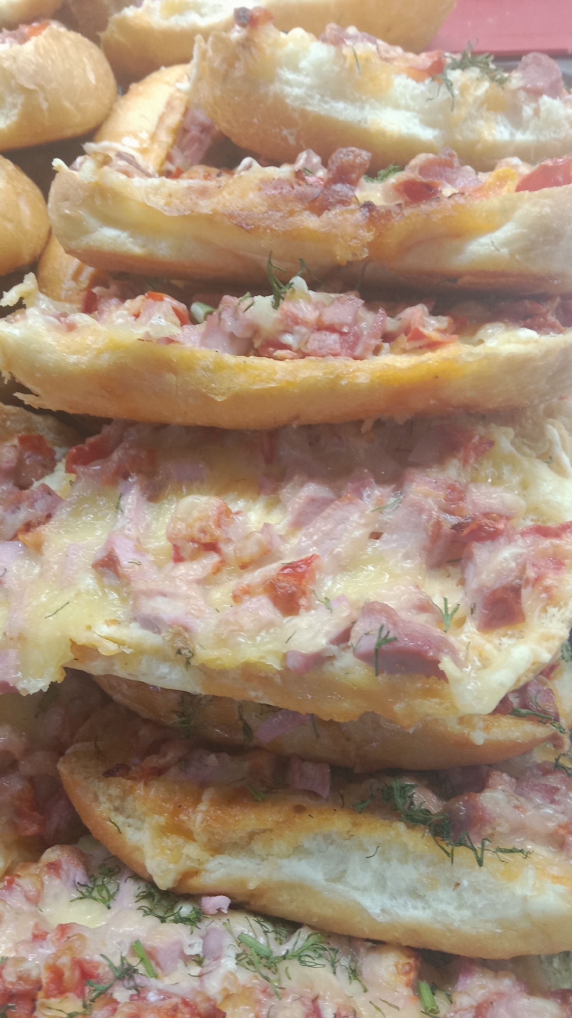 The image shows a close-up of several open-faced sandwiches or mini-pizzas stacked on top of each other. The bread appears to be a baguette or similar long roll, split lengthwise and topped with melted cheese, chopped ham or sausage, and possibly some tomatoes and herbs like dill. The sandwiches are golden brown and appear to be freshly baked. The focus is on the food items, and the background is blurred, suggesting this might be a food display or a personal food photograph. There are no people, actions, or environmental elements identifiable in the image that would indicate a specific location in Petropavl, Kazakhstan. The scene is purely focused on the food.