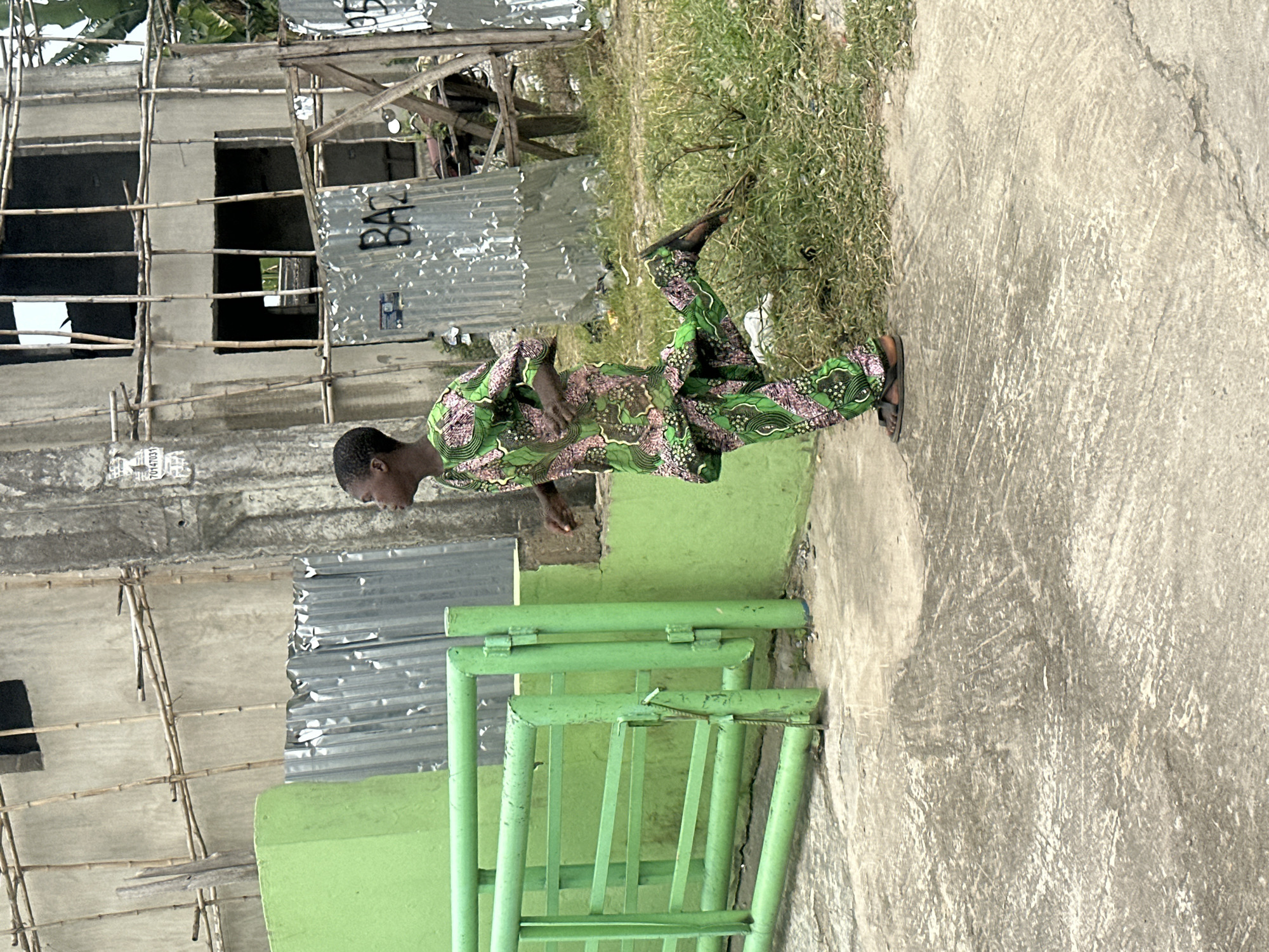 A young Black person, wearing a vibrant green and pink patterned outfit and dark sandals, is captured mid-stride while running. The individual is facing right, with their right leg extended backward and their left leg forward, suggesting forward motion. They are running on a rough concrete surface, adjacent to a patch of dry, sparse grass.
Behind the running figure, a bright green metal gate with horizontal bars and vertical posts stands partially open. A light green wall is visible behind the gate. Further back, a building under construction is apparent, with concrete walls and bamboo scaffolding supporting its unfinished structure. Several sheets of corrugated metal are leaning against the building. One sheet has the word "BAZ" scrawled on it. The environment appears to be an urban setting, possibly a developing area, given the construction and materials visible. The overall impression is one of dynamic movement within a somewhat dilapidated urban landscape. The image was taken in Badagry, Nigeria.