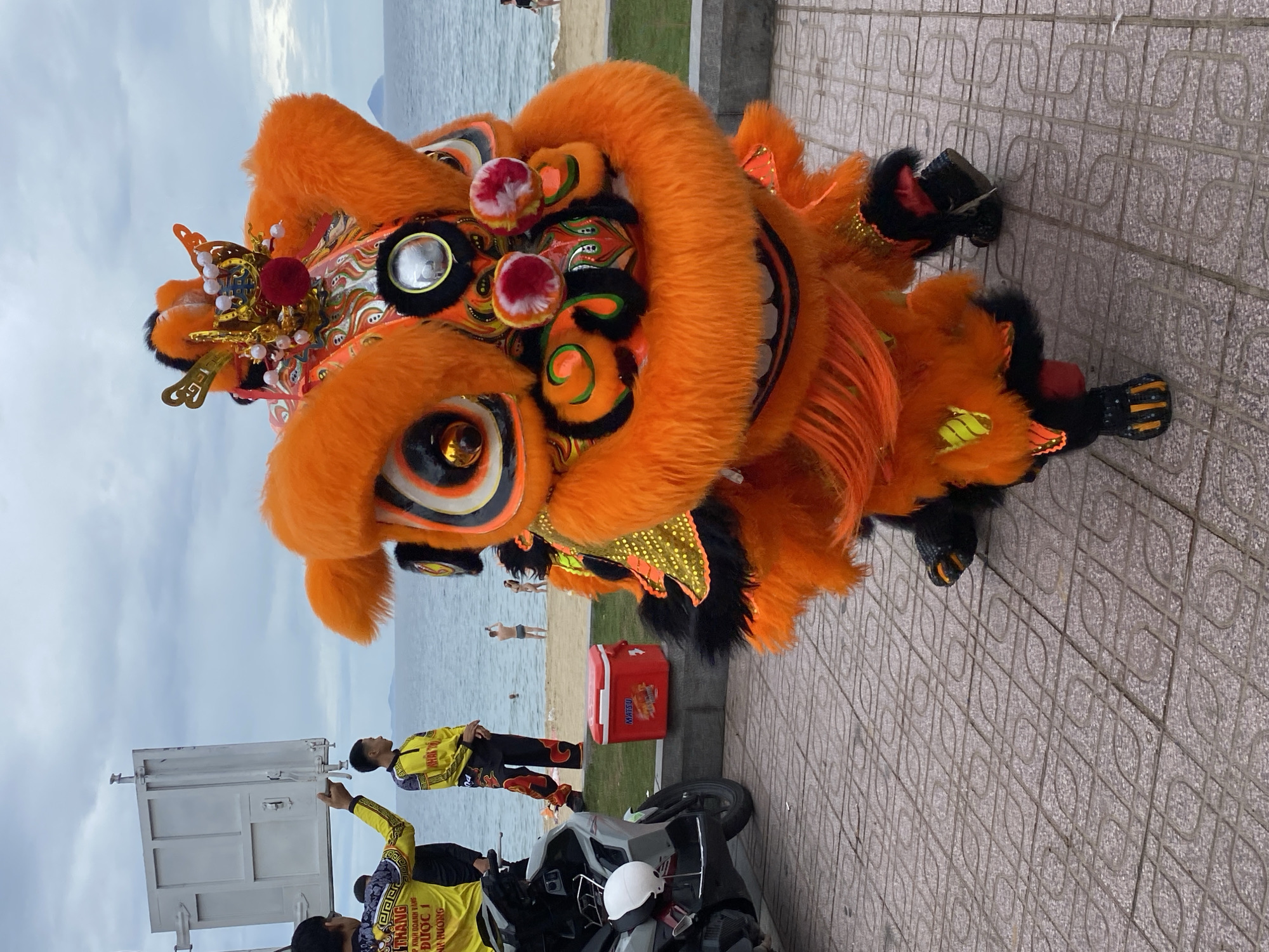 This is a medium shot, eye-level, outdoor photograph taken on a cloudy day in Nha Trang, Vietnam. The primary subject is a vibrant, orange lion dance costume with large, expressive eyes and a gaping mouth, positioned in the foreground. The costume has fur and decorative elements. Behind the lion, a tiled walkway leads to a beach area. Several people are present: two men in yellow shirts and black pants are visible on the left, one holding a white rectangular object. Further back, on the beach, a few individuals are seen, some in swimwear. A red cooler with the brand name "MATSU" sits on a concrete ledge to the right of the walkway. A white motorcycle is parked on the left side of the image. The sea and the distant coastline are visible in the background.
