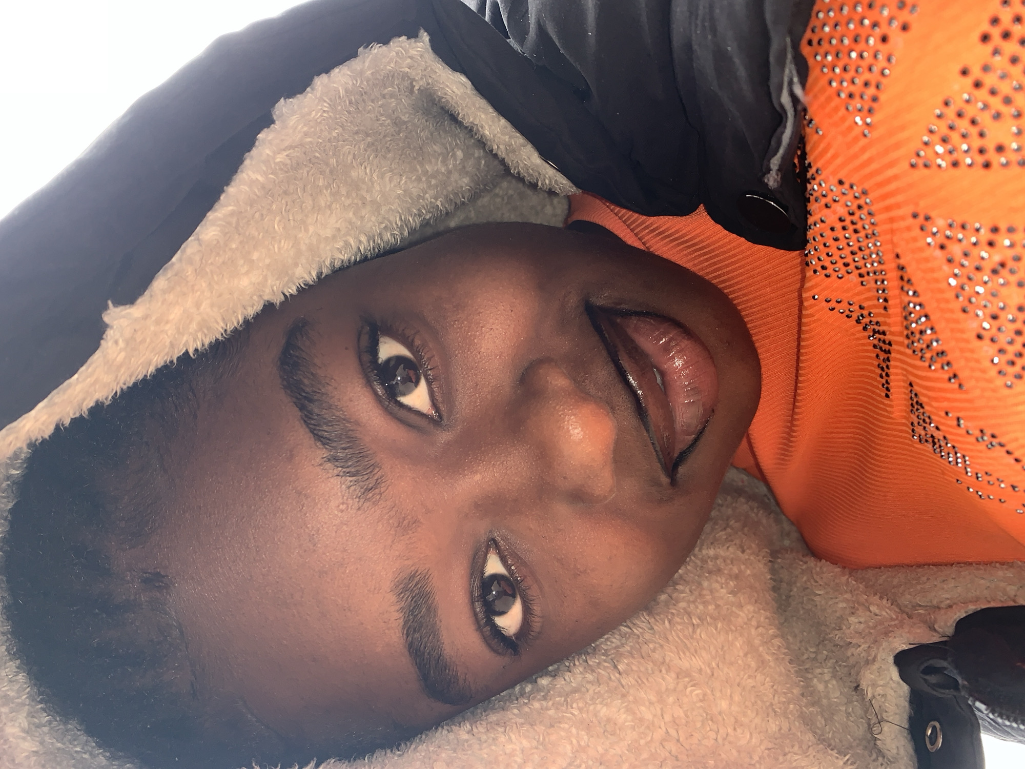 A close-up, low-angle shot features a young Black person wearing a hooded jacket and an orange top adorned with rhinestones. The person's face is visible, with eyes looking forward and a slight smile. The jacket's fuzzy lining frames the face. The setting is outdoors, with a bright, overexposed sky in the background. The image is taken in Rafin Par, Nigeria.