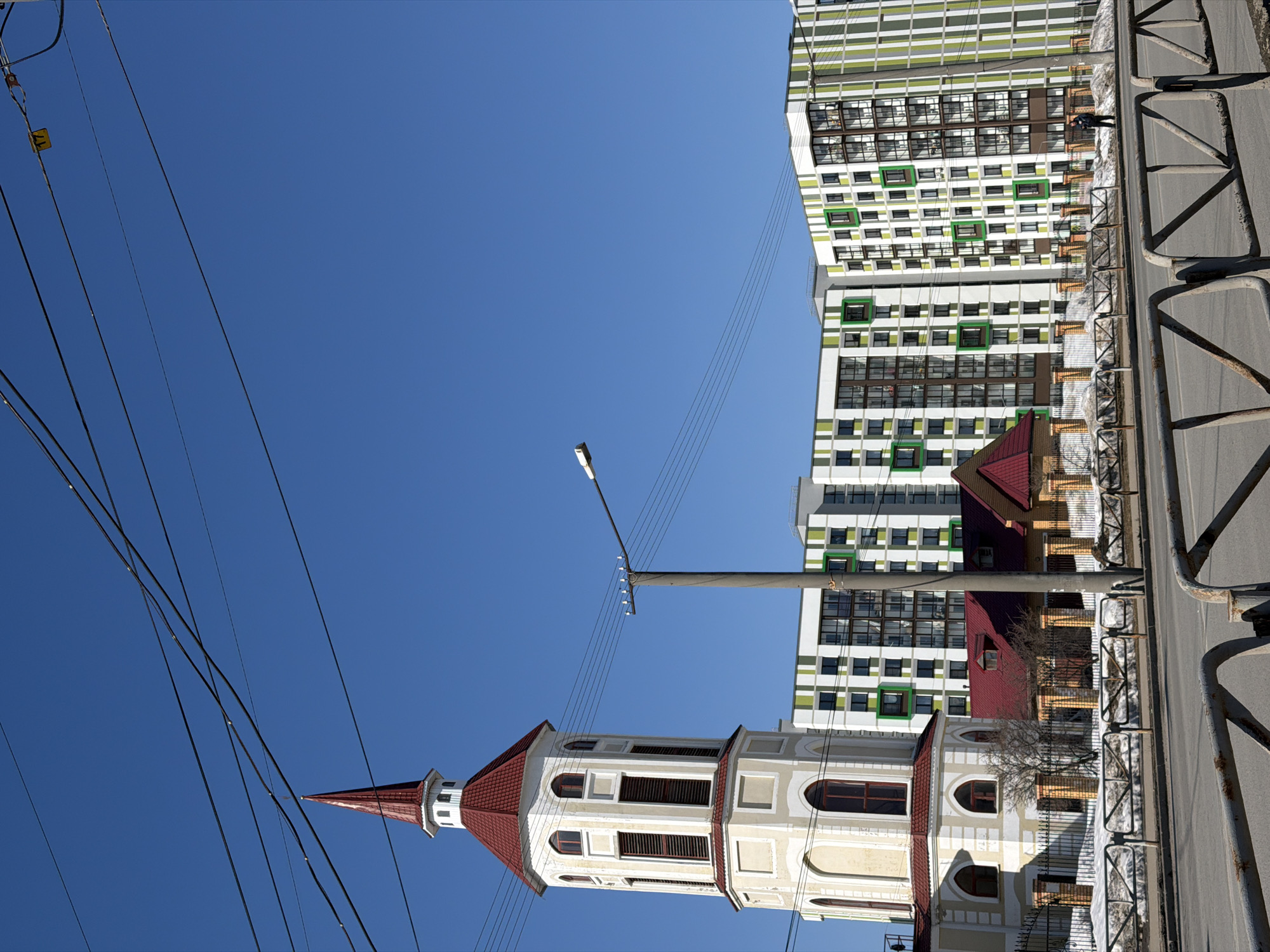 The image, likely taken in Berezniki, Russia, presents a view of an architectural juxtaposition. In the foreground stands a light-colored, multi-story bell tower, topped with a red conical roof and a spire. The tower features vertical windows with dark shutters or coverings. Overhead power lines intersect the view of the tower against a clear blue sky. Adjacent to the tower, on the right side of the frame, rises a tall, modern apartment building. The building has a white facade with numerous windows, some outlined in green. A smaller structure with a red roof is nestled between the tower and the apartment building. A road runs alongside the tower and buildings, with a metal guardrail visible on the edge of the roadway. A person is visible on the sidewalk in front of the apartment building.