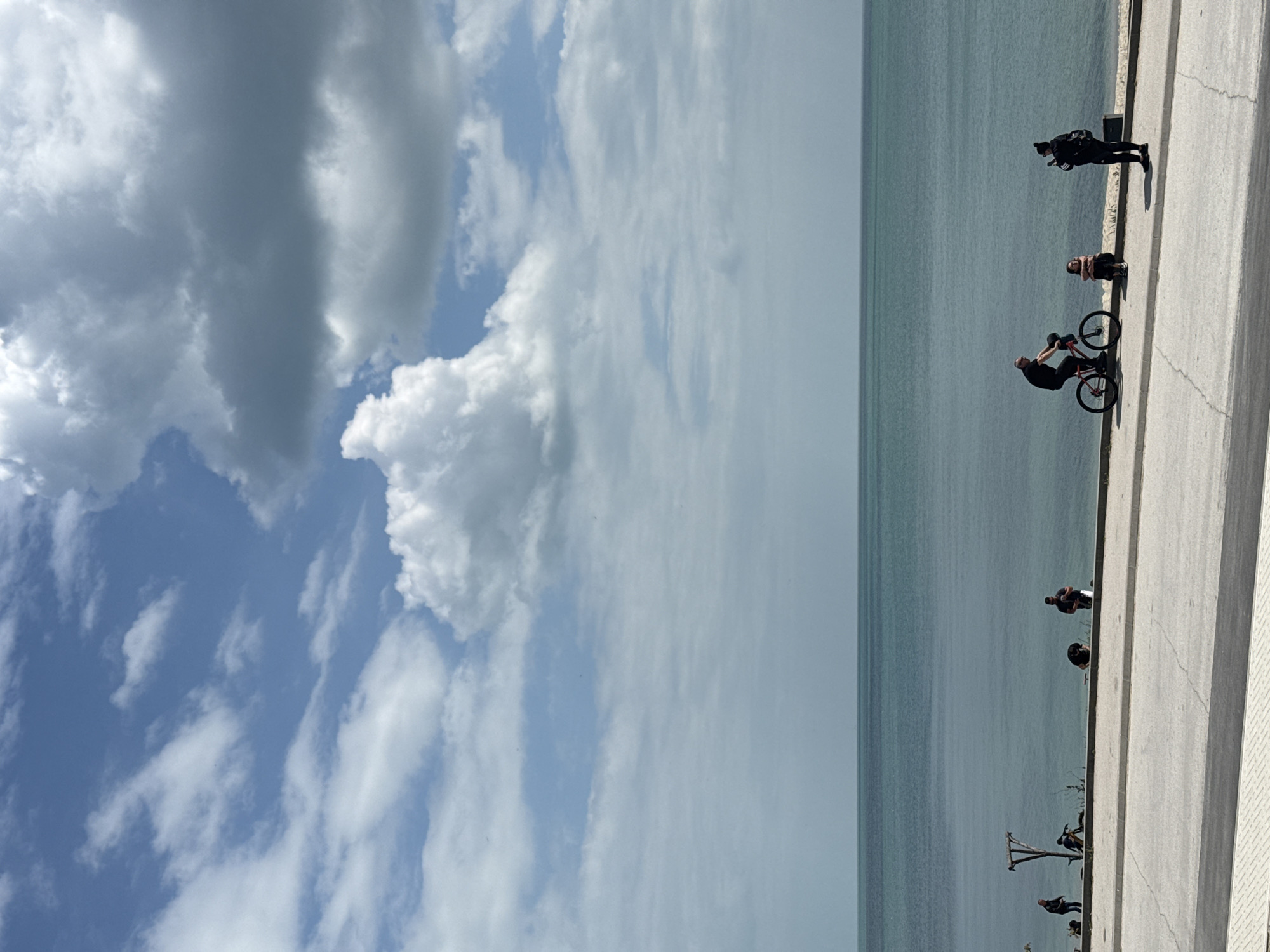 The image shows a waterfront scene in Varna, Bulgaria. On the right, a concrete promenade borders the sea. Several people are visible along the promenade: one person is standing and taking a photo. Further along, another is sitting next to a person with a bicycle. Other individuals are scattered along the promenade, some sitting. To the left, the sea extends to the horizon, with the sky filled with clouds. The image appears to have been taken at midday during calm weather.