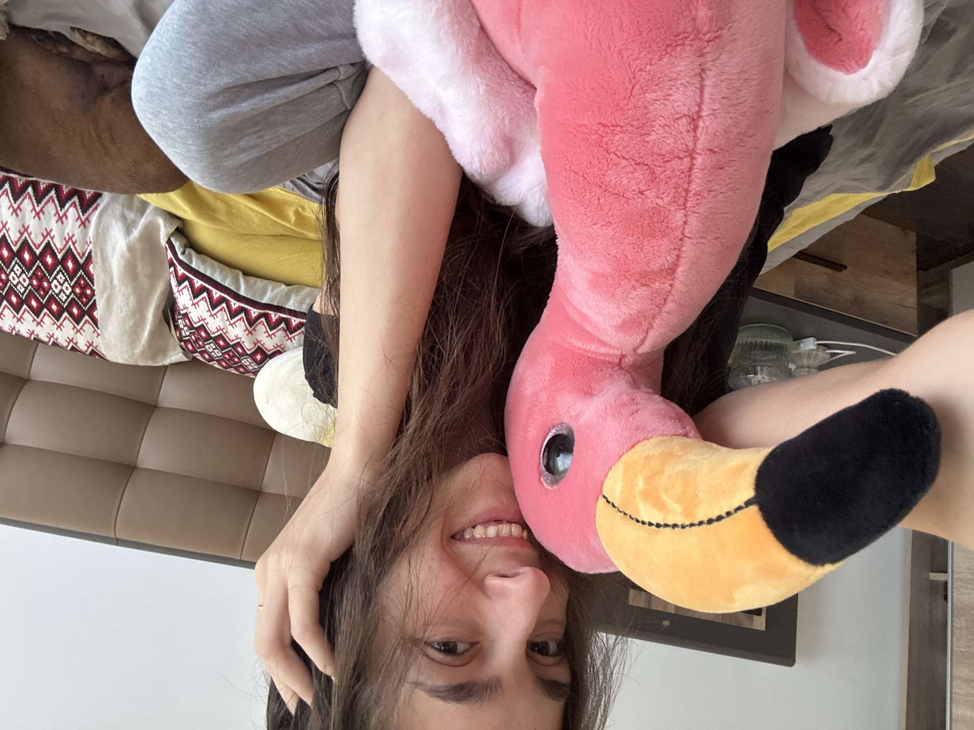 A woman with dark hair is lying upside down on a bed, smiling at the camera, with a large pink flamingo plush toy nestled against her face. The bed is covered with various pillows and blankets. The room appears to be indoors, possibly a bedroom. There is no information in the image to indicate this photo was taken in Sofia, Bulgaria.