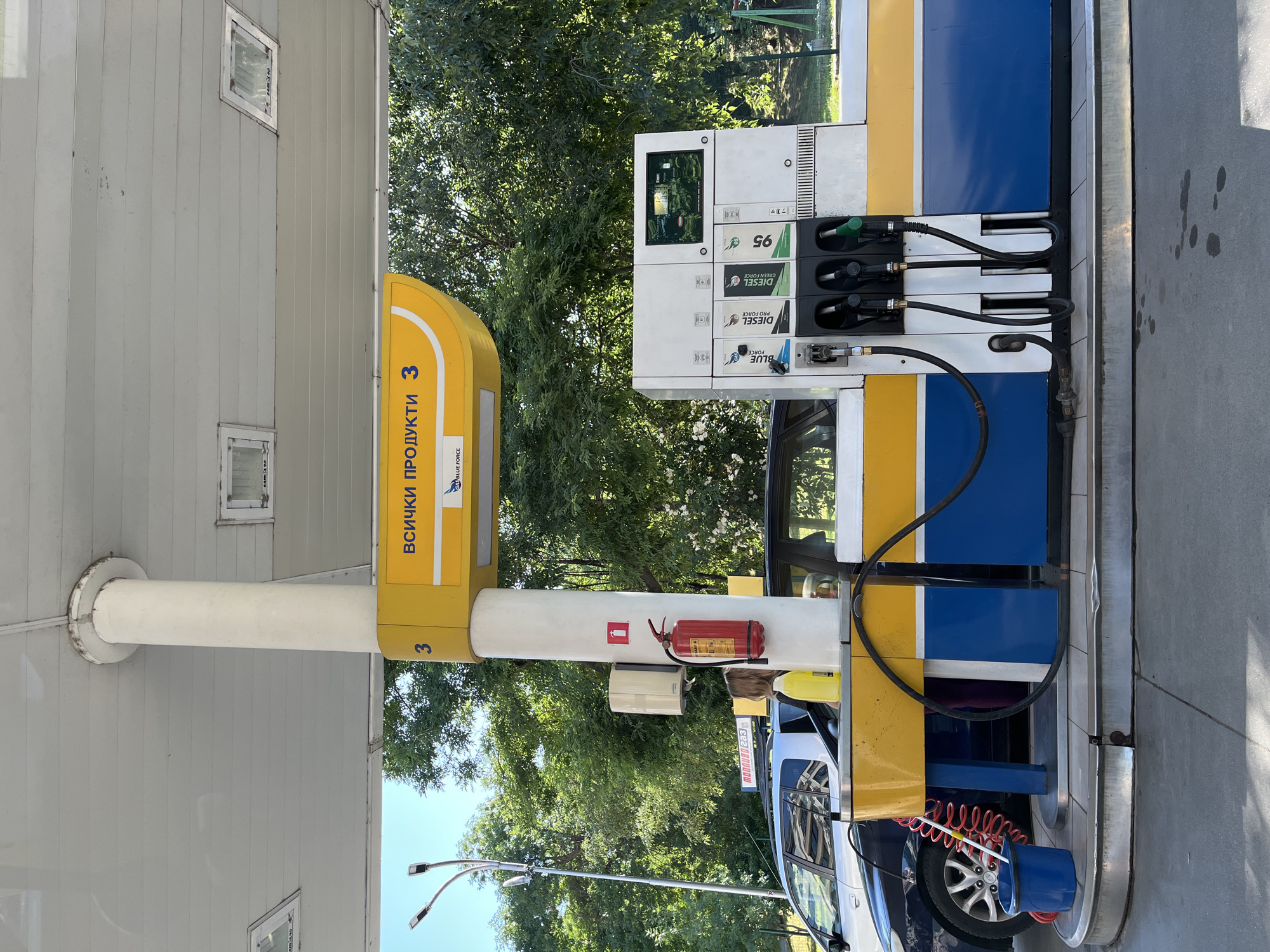 The image shows a gas station with a prominent yellow and blue color scheme. A white pillar supporting the canopy has a yellow sign with the Cyrillic text "ВСИЧКИ ПРОДУКТИ 3" which translates to "ALL PRODUCTS 3" and the logo "GAS BLUE FORCE". On the pillar, to the left of the sign, is a red fire extinguisher and a white dispenser. A car is parked at the pump, and the fuel dispenser unit features multiple nozzles and a screen displaying fuel prices, with labels indicating "BLUE FORCE", "DIESEL", and "95". A red coiled hose and a blue bucket are visible on the ground near the pump. The background consists of lush green trees and a bright blue sky, suggesting an outdoor urban setting. Given the "Blue Force" branding, this is likely a Blue Force gas station. A sign on the car reads "МОБИЛОВ 233", which could be a phone number or identification code. This image appears to be taken in Plovdiv, Bulgaria, a major city known for its gas stations.