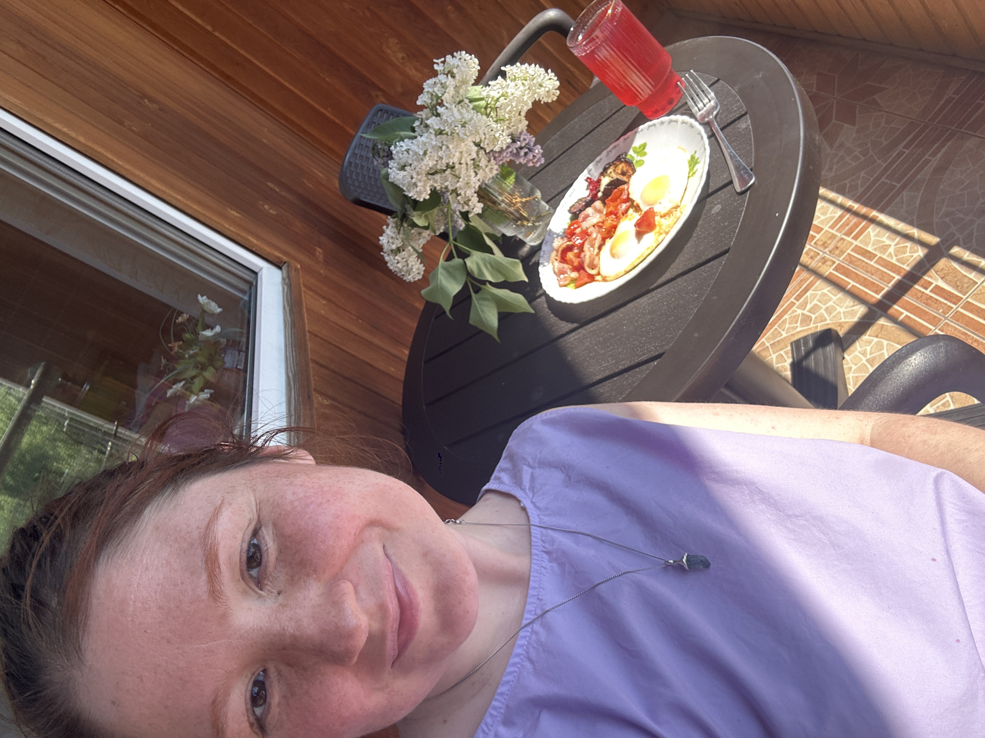 A selfie taken in an outdoor setting, likely a patio or deck. The person taking the photo is a fair-skinned individual with reddish-brown hair, wearing a light purple shirt and a necklace. They are smiling slightly at the camera.
To the right of the person, there is a small, round black table. On the table, there is a plate of food that appears to be a breakfast dish with fried eggs, bacon, and some vegetables, along with a fork. Beside the plate, there is a glass filled with a red liquid. There is also a small vase holding white flowers, and a glimpse of the exterior of a building with wood paneling and a window reflecting the surrounding greenery. The surface visible in the background to the right looks like patterned tiles.
There is no information to suggest that this image was taken in the city of Nizhny Novgorod, Russia.