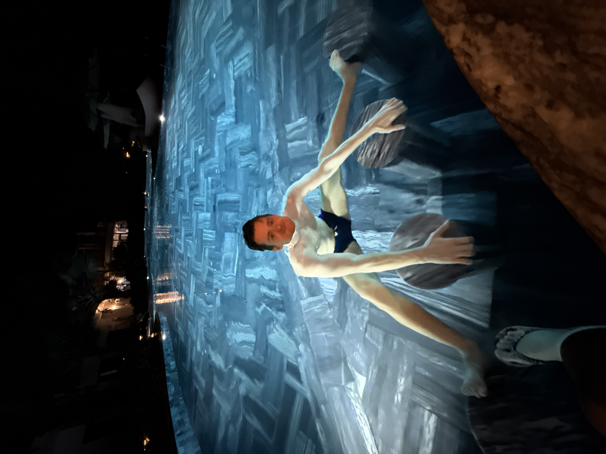 The image depicts a nighttime scene in a swimming pool, likely at a resort in Phú Quốc, Vietnam. The central subject is a male figure, partially submerged in the blue-grey water. His face, which appears to be superimposed, shows a slight smile. He is wearing dark swim trunks, and his arms and legs are visible above the water, seemingly in a relaxed or floating pose.
The pool features several large, dark, circular objects that resemble stepping stones or decorative elements. In the bottom right, a foot wearing a light-colored slipper is visible, suggesting another person is nearby or taking the photo. A rough, brown-textured object, possibly a natural rock formation or wooden structure, occupies the upper right foreground.
The background is dark, indicating it is night, with distant buildings and vegetation dimly illuminated by artificial lights, reflecting on the water. The water itself has a distinctive patterned tile design. The overall atmosphere suggests leisure and enjoyment in a resort setting. No text is visible in the image. The image is oriented sideways.