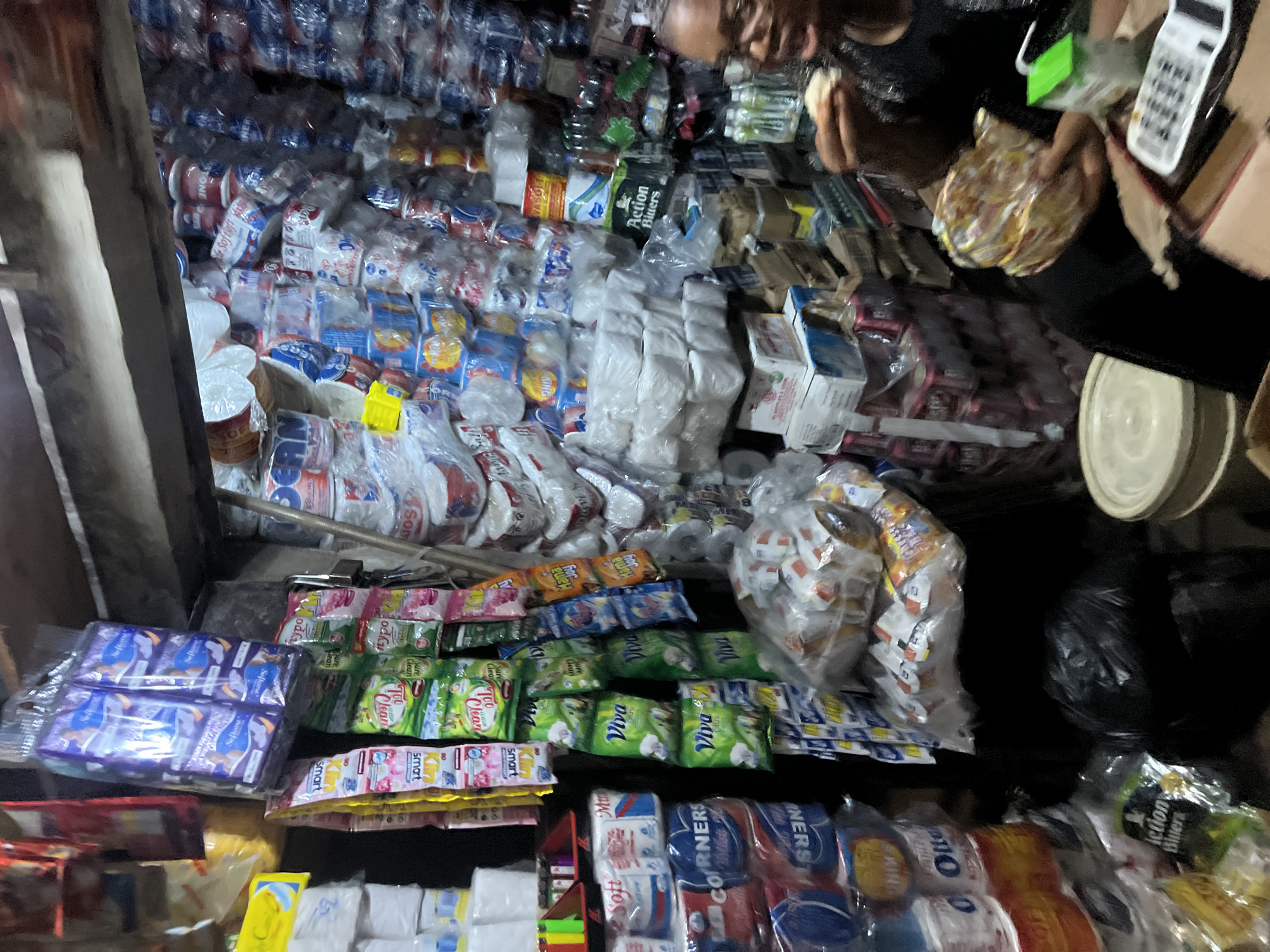 The image depicts a densely packed small shop, likely a convenience store or market stall, in Nkpor, Nigeria. The shop is filled from floor to ceiling with various packaged goods, including toiletries, food items, and beverages. Stacks of toilet paper rolls, likely branded "Corners" and "Viva," are prominently displayed. Bags of what appear to be sanitary pads, possibly "Softex," hang from the ceiling. Rows of canned drinks and bottled beverages are also visible. In the foreground, a woman is seated and appears to be eating or inspecting a food item. Another individual is partially visible in the background, also engaged in an activity. The lighting suggests an indoor environment, possibly with limited natural light. The overall impression is one of a busy, well-stocked local market.