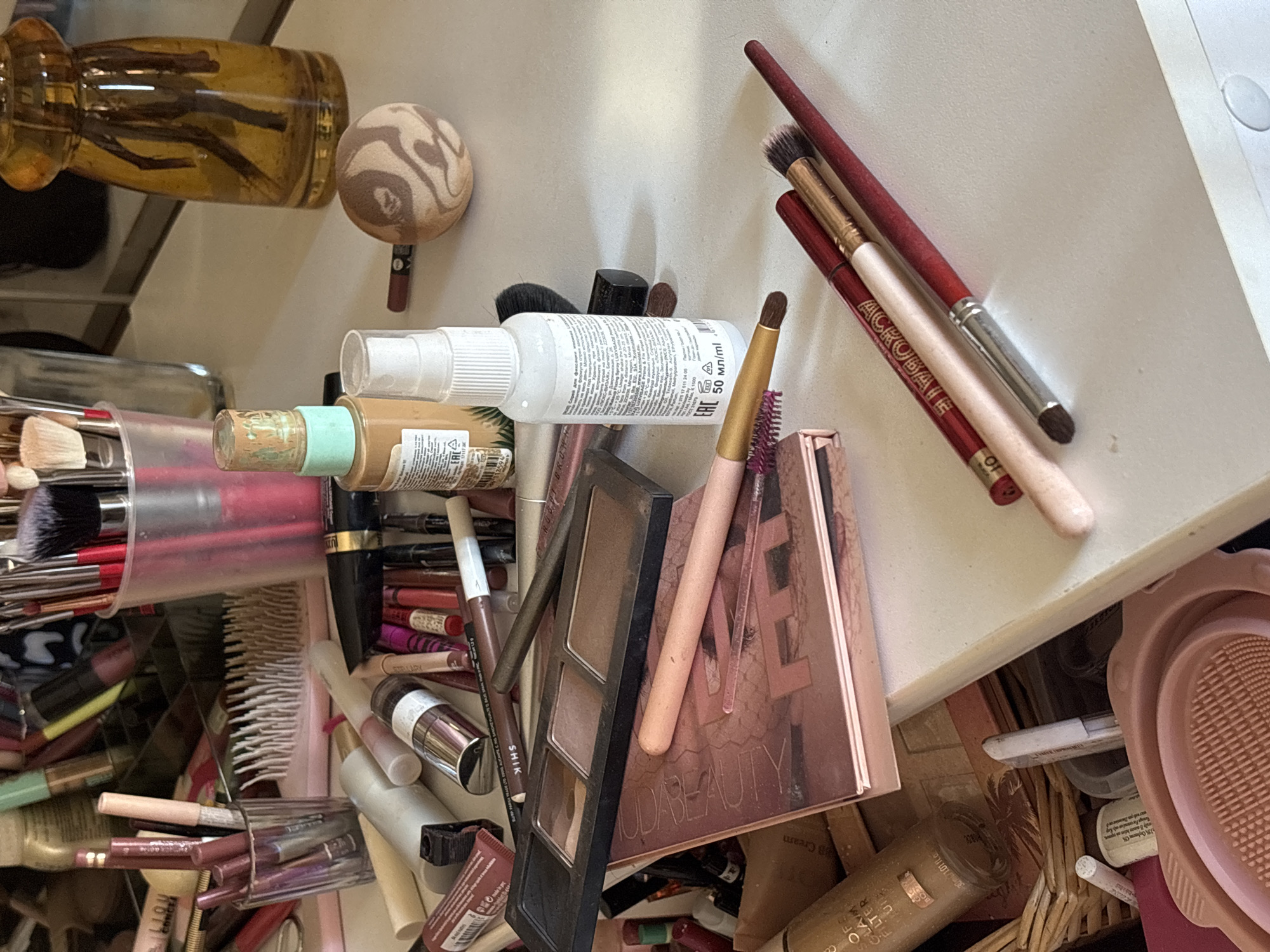 A top-down view depicts a cluttered makeup drawer. The items include various makeup brushes, palettes, spray bottles, foundation, lipsticks, and other cosmetic products. Text is visible on several products, identifying brands like "HUDA BEAUTY", "SHIK", "ACROBATE", and "STELLARY". The chaotic arrangement suggests a lack of organization within the drawer.