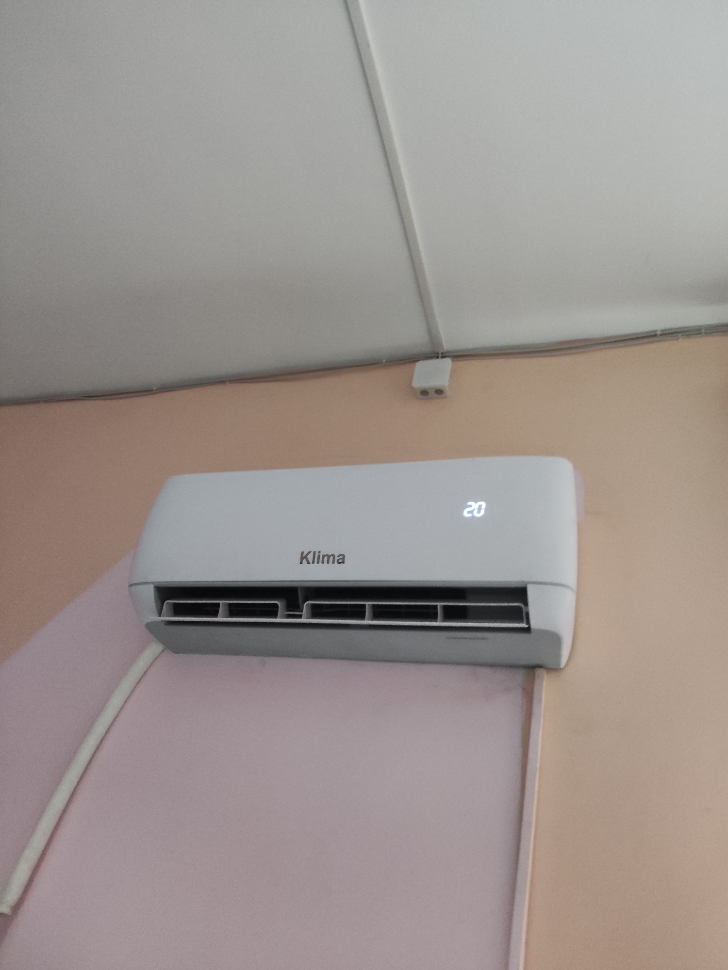 A white air conditioner is mounted high on a wall. The air conditioner has a digital display showing "20". The brand name "Klima" is visible on the front of the unit. A white cable runs from the air conditioner down the wall. The wall appears to be painted a peach color, and the ceiling is white. This image was taken in Shymkent, Kazakhstan.