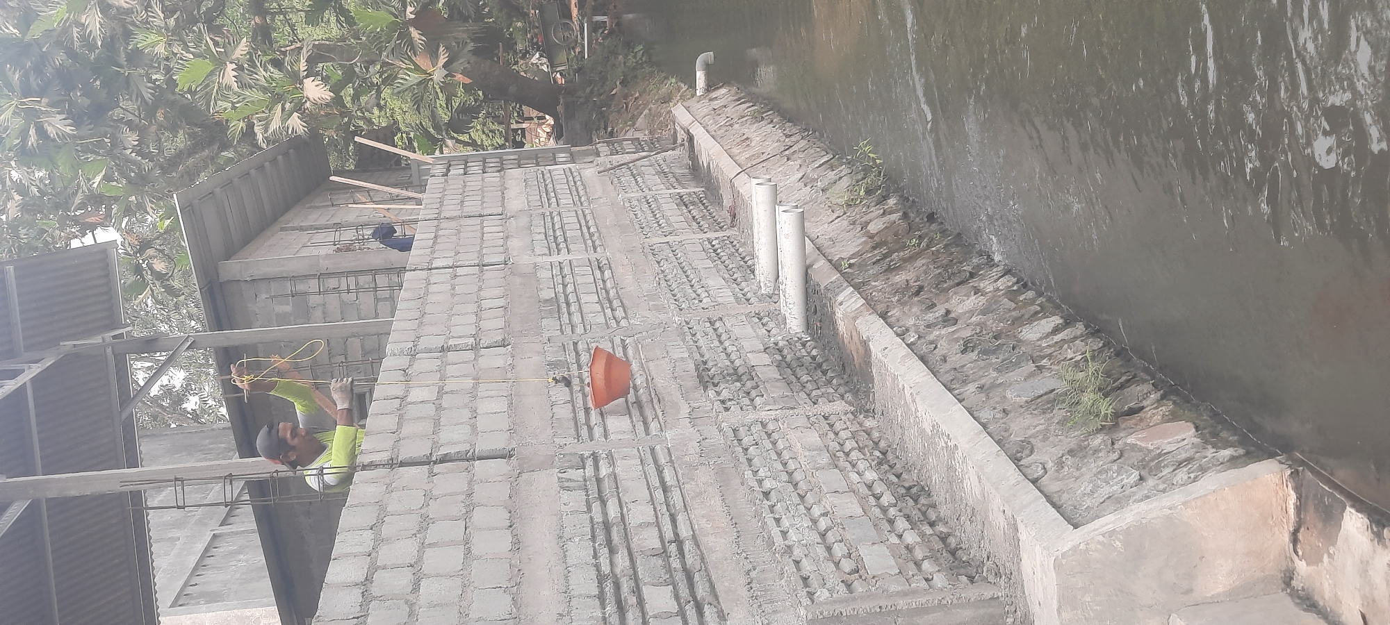 In the city of Ploso, Indonesia, construction is underway next to a body of water. A man wearing a bright yellow shirt and a cap is working on the upper level of a brick wall structure, holding a yellow rope. Another construction worker, wearing a blue shirt, is visible in the background, also on the upper level. An orange bucket is suspended by a rope from the wall. The building appears to be in the early stages of construction, with exposed rebar and unfinished brickwork. There are two white pipes protruding from the wall, extending over the water. The water has a slightly murky appearance and shows some ripples. Lush green trees and foliage are visible behind the construction site, indicating a natural environment surrounding the urban area. The overall scene depicts active construction in a semi-natural setting.