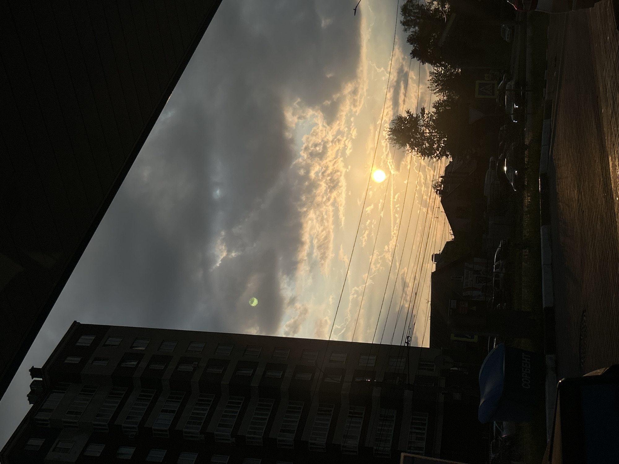 The image captures a sunset scene in the city of Ivanovo, Russia. A tall, multi-story apartment building stands on the left side of the frame, its facade dark against the bright sky. The sky is filled with dramatic clouds, illuminated by the setting sun, which appears as a bright, circular orb in the middle right of the image. Wires crisscross the sky in the foreground. Below the sky and buildings, a street or parking area is visible, paved with brickwork, with several cars parked or driving. A blue trash bin with Russian text is located on the right side of the apartment building. The overall impression is of an urban environment at dusk, with the natural beauty of the sunset contrasting with the man-made structures.