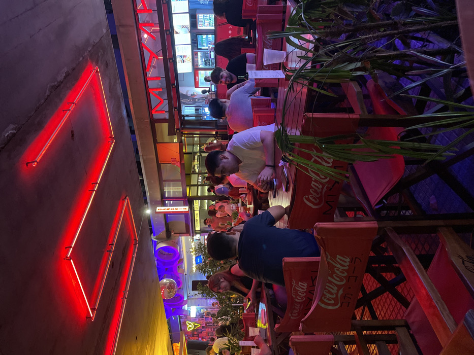 An outdoor restaurant, lined with red Coca-Cola-branded chairs and tables, is brightly lit. A neon sign resembling a slanted square is on the wall. People are seated at the tables. In the background, signs and decorations hang from the ceiling. The lighting shifts from red to purple and blue. Some greenery hangs over the tables.