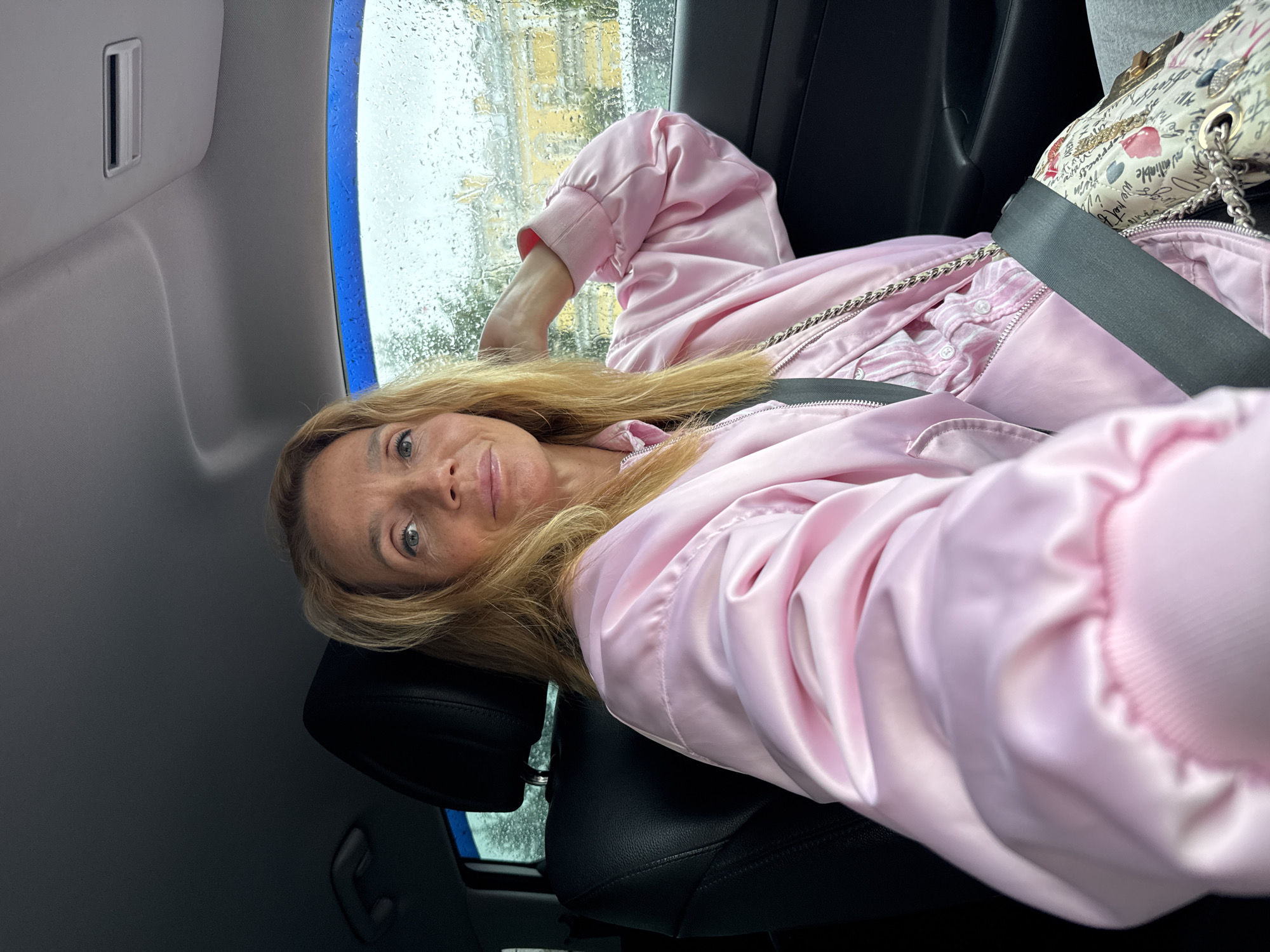 A woman with blonde hair is seated in the passenger seat of a car, wearing a pink satin bomber jacket over a pink and white striped shirt. She has her right hand behind her head, and she is looking at the camera with a slight smile. The car's interior is visible, including the black leather seat and the dashboard. Through the rain-streaked window, a yellow building with arched windows can be seen, suggesting the location is the city of Saint Petersburg, Russia. The overall mood is casual and relaxed.